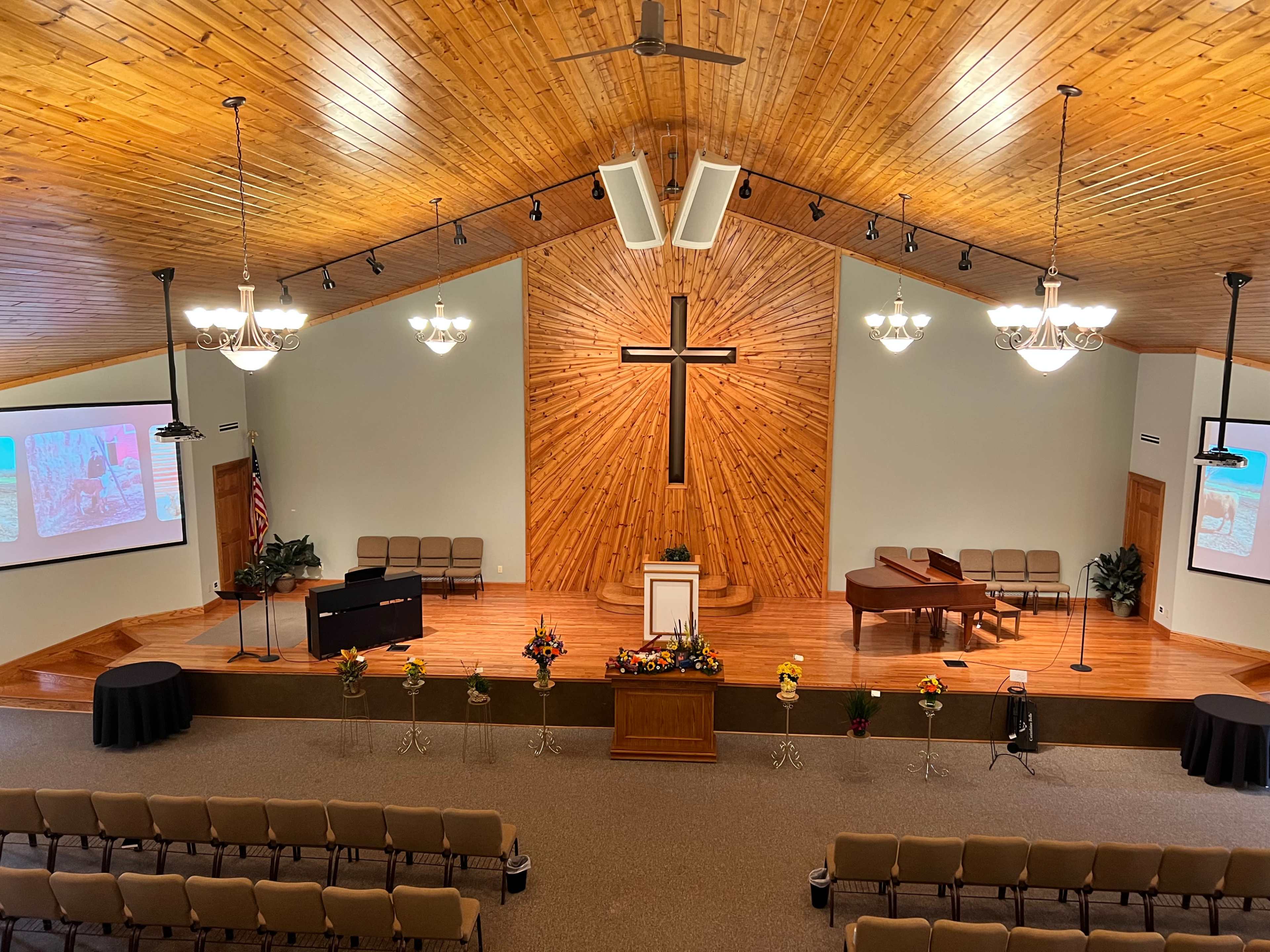 The interior of a church features wooden paneling, a large cross on the wall, a stage with a piano, and rows of chairs arranged for a congregation.