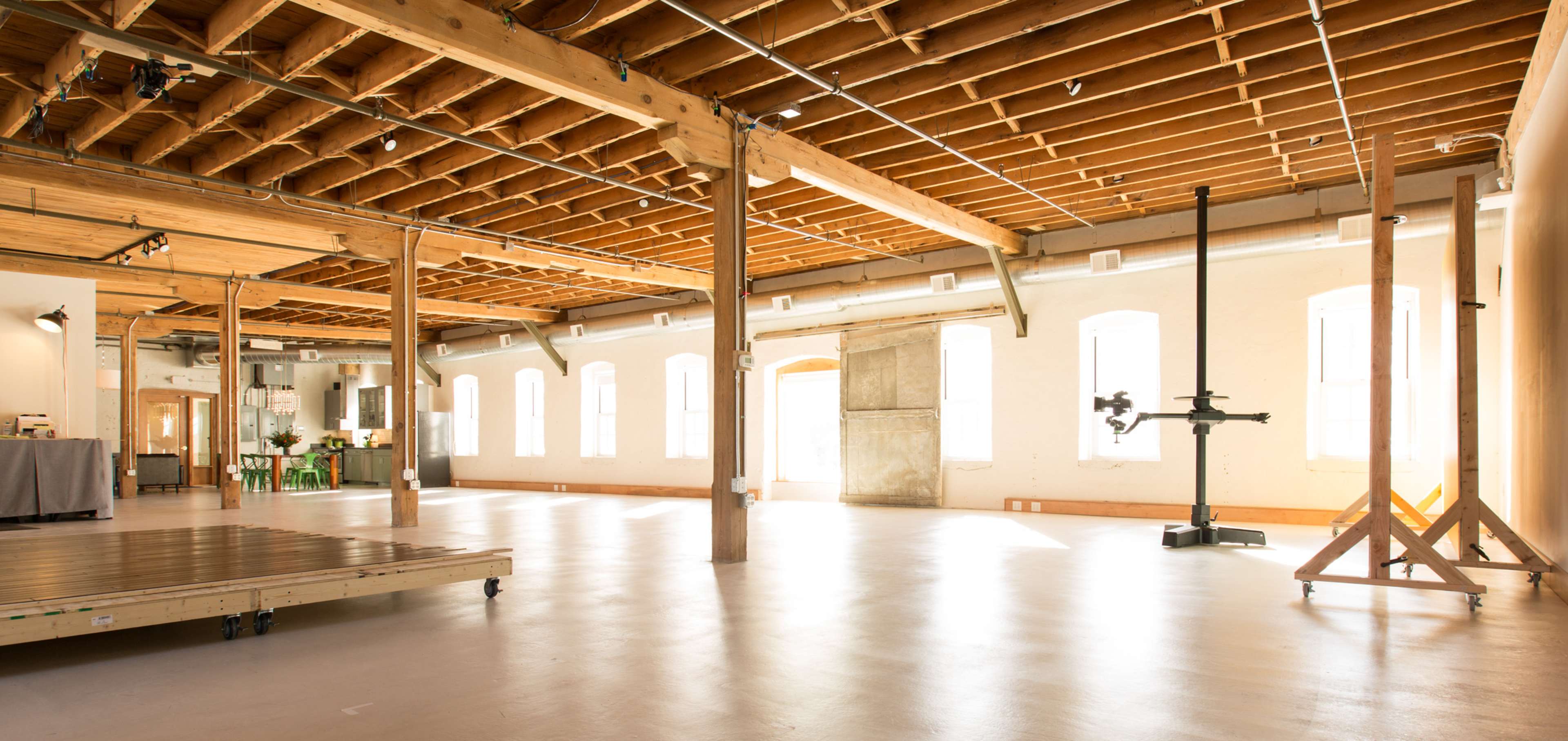 The image depicts an open, well-lit loft space with wooden beams and a polished floor, featuring large windows and minimal furniture.