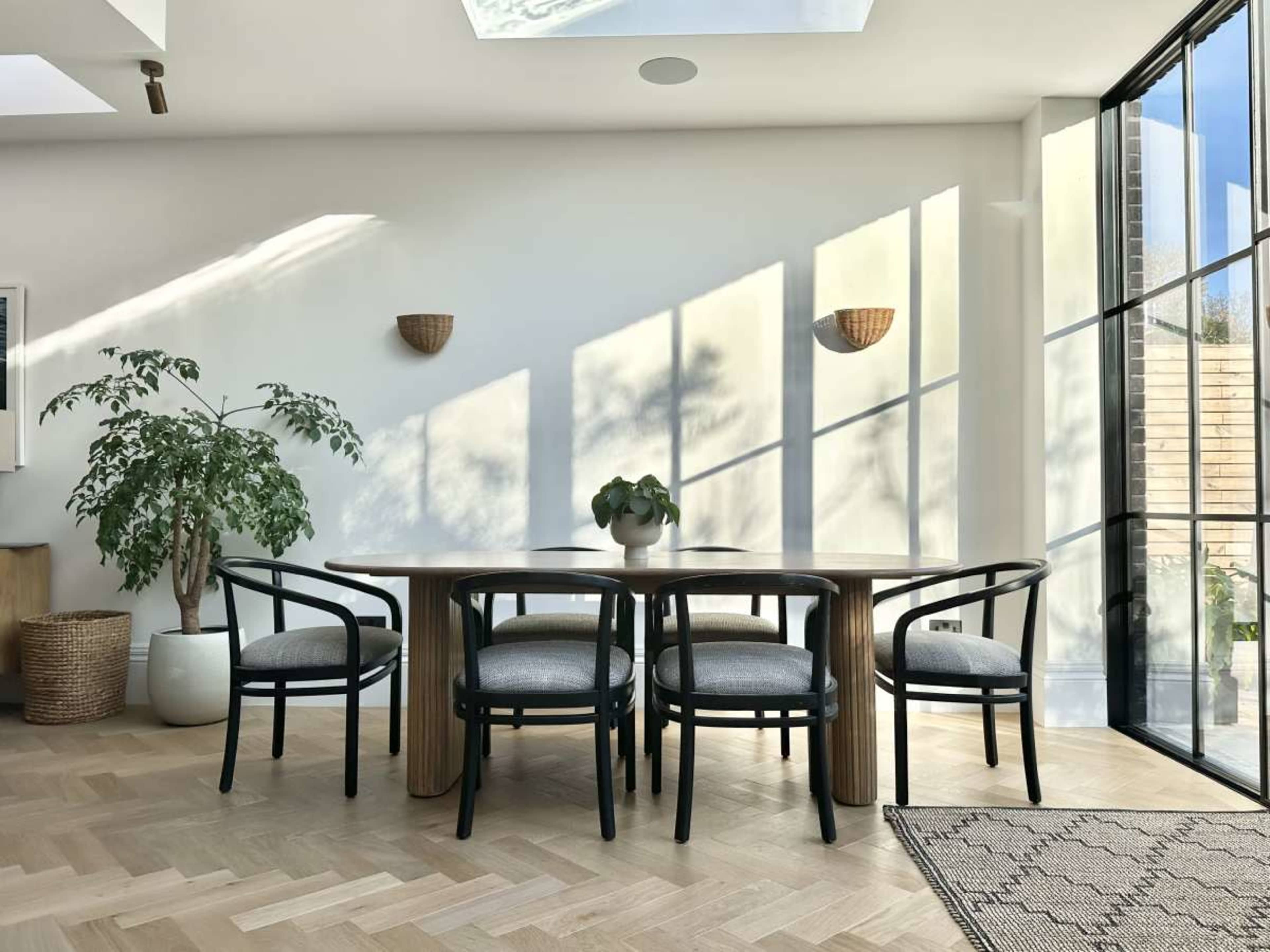 A dining area features a wooden table surrounded by four black chairs, with a large window allowing natural light to cast shadows on the floor.