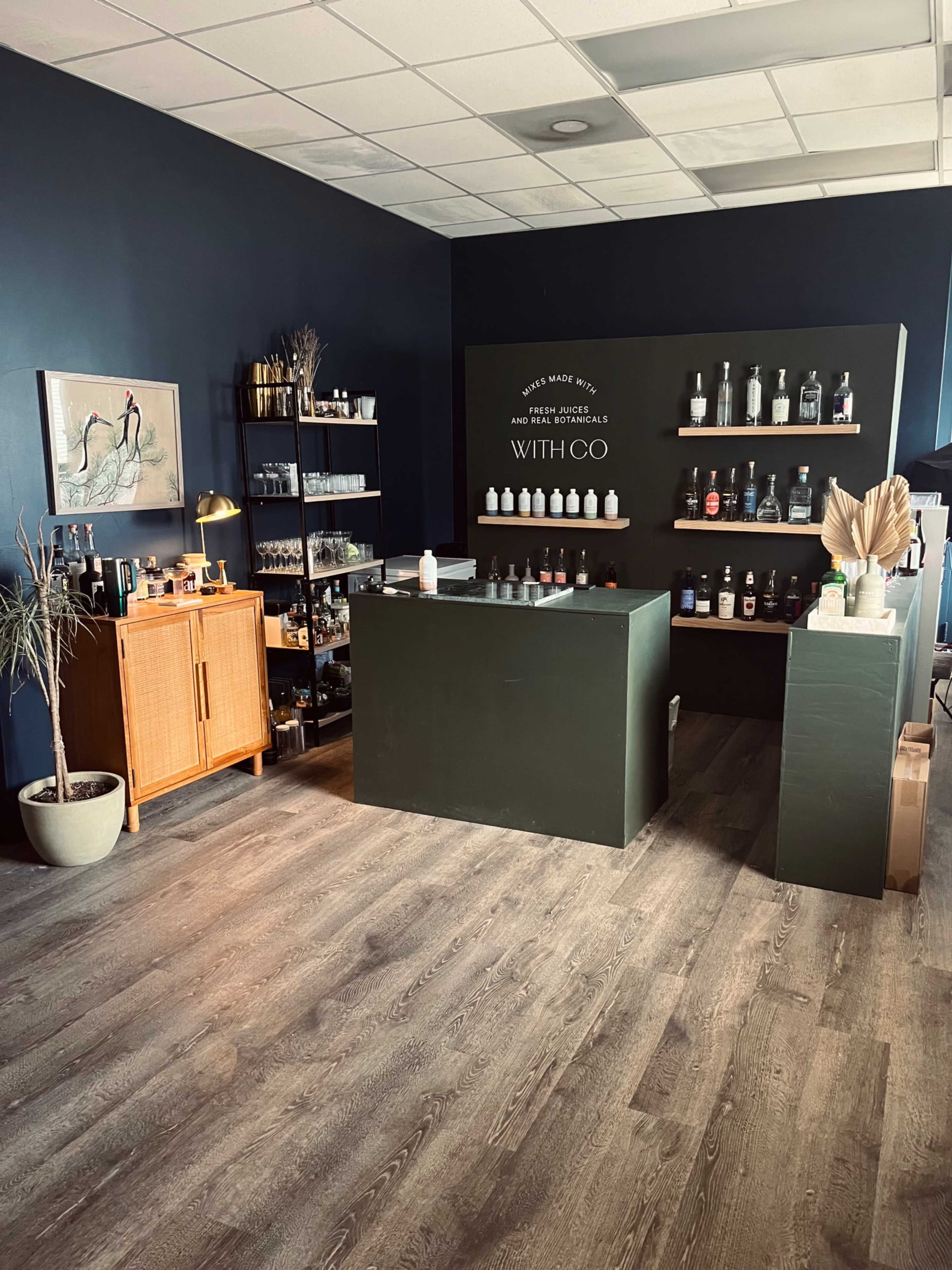 The image shows a modern retail space featuring a dark blue wall, shelves stocked with various bottles, a wooden countertop, and a display of plants and decor items.