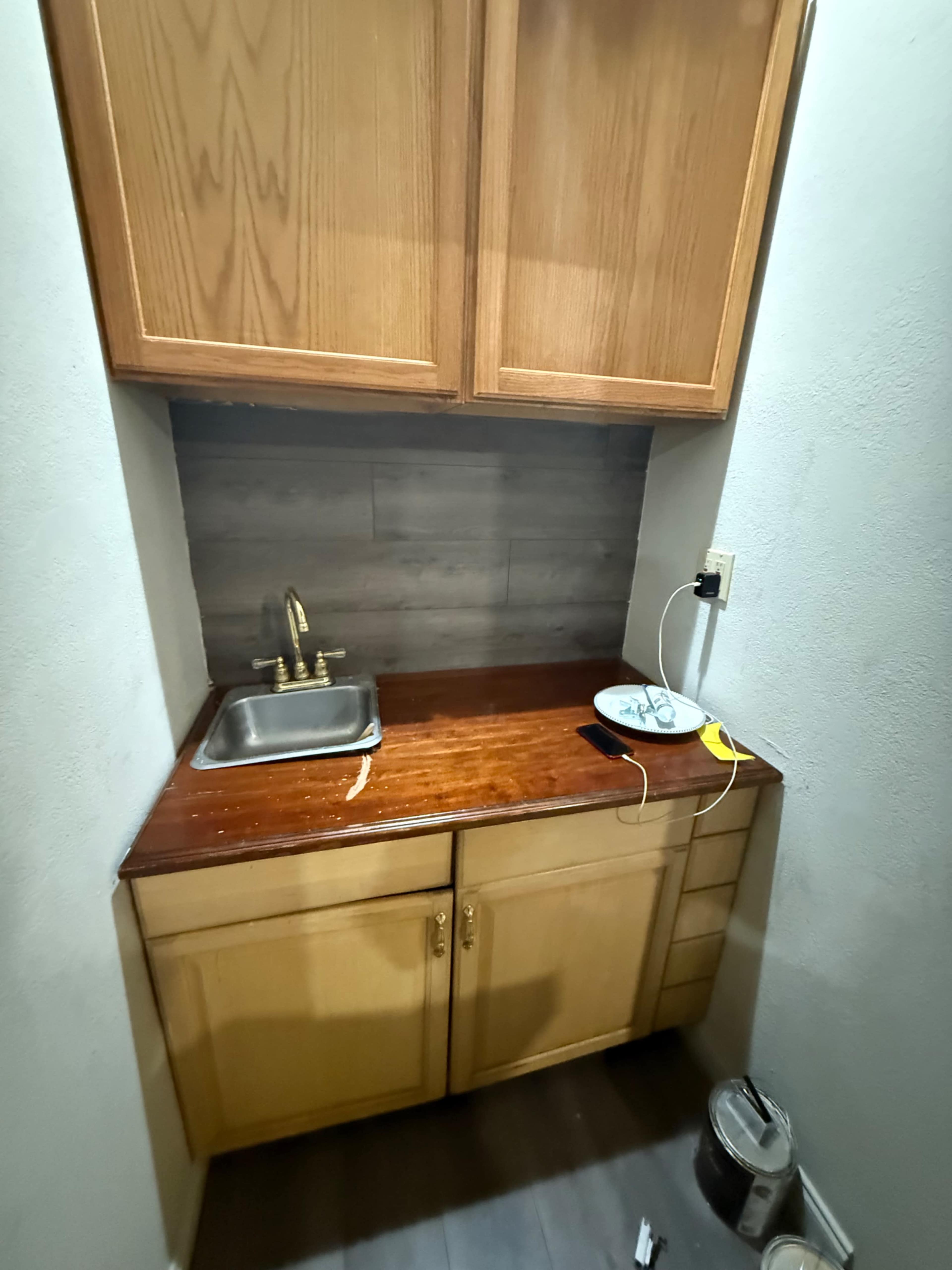 The image shows a small kitchenette area with a sink, wooden cabinets above and below, and a countertop with a plate and a charging phone.