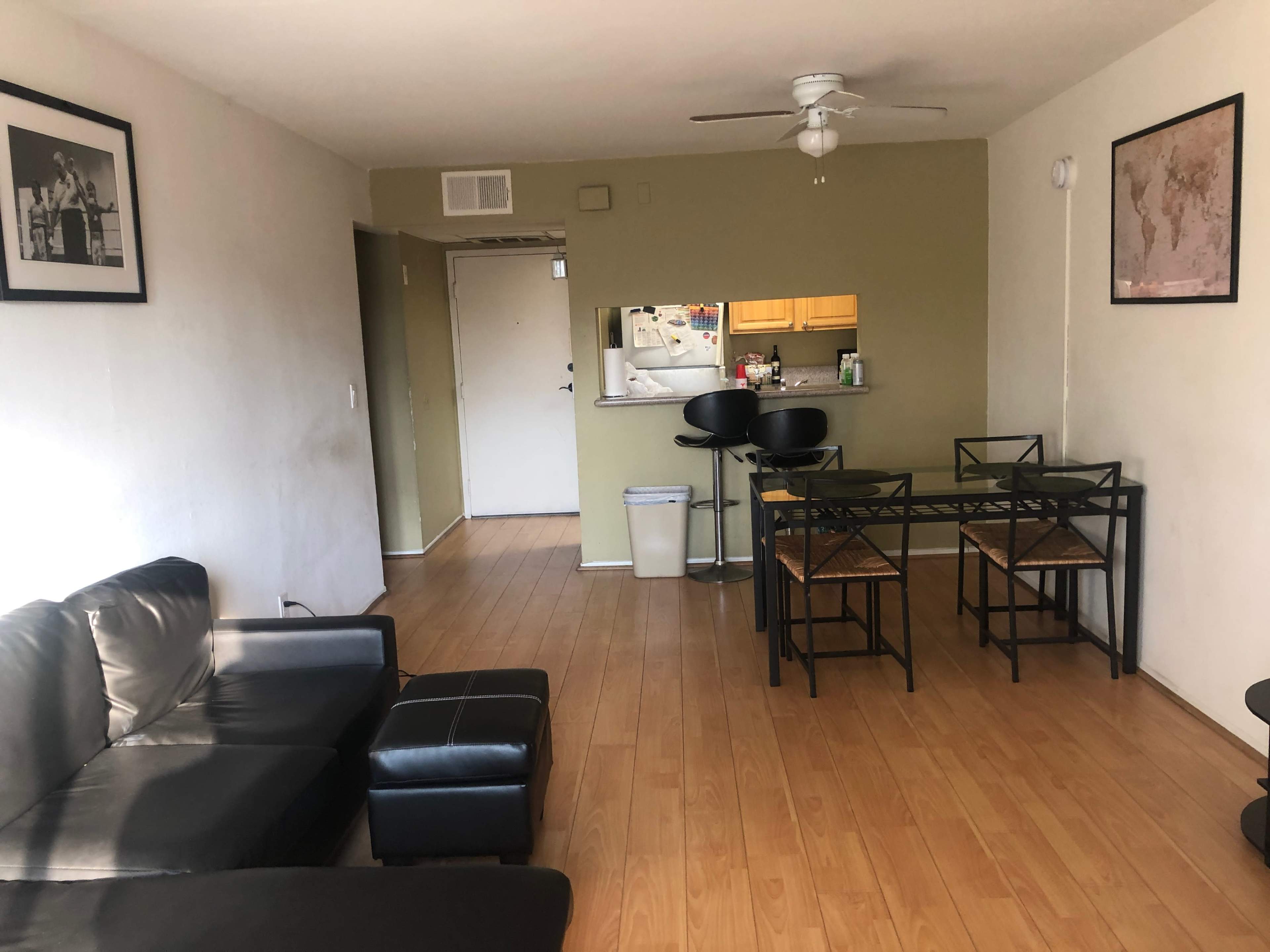 A living space with a black sofa, a matching ottoman, a glass dining table with four chairs, and a view of a kitchen area in the background.