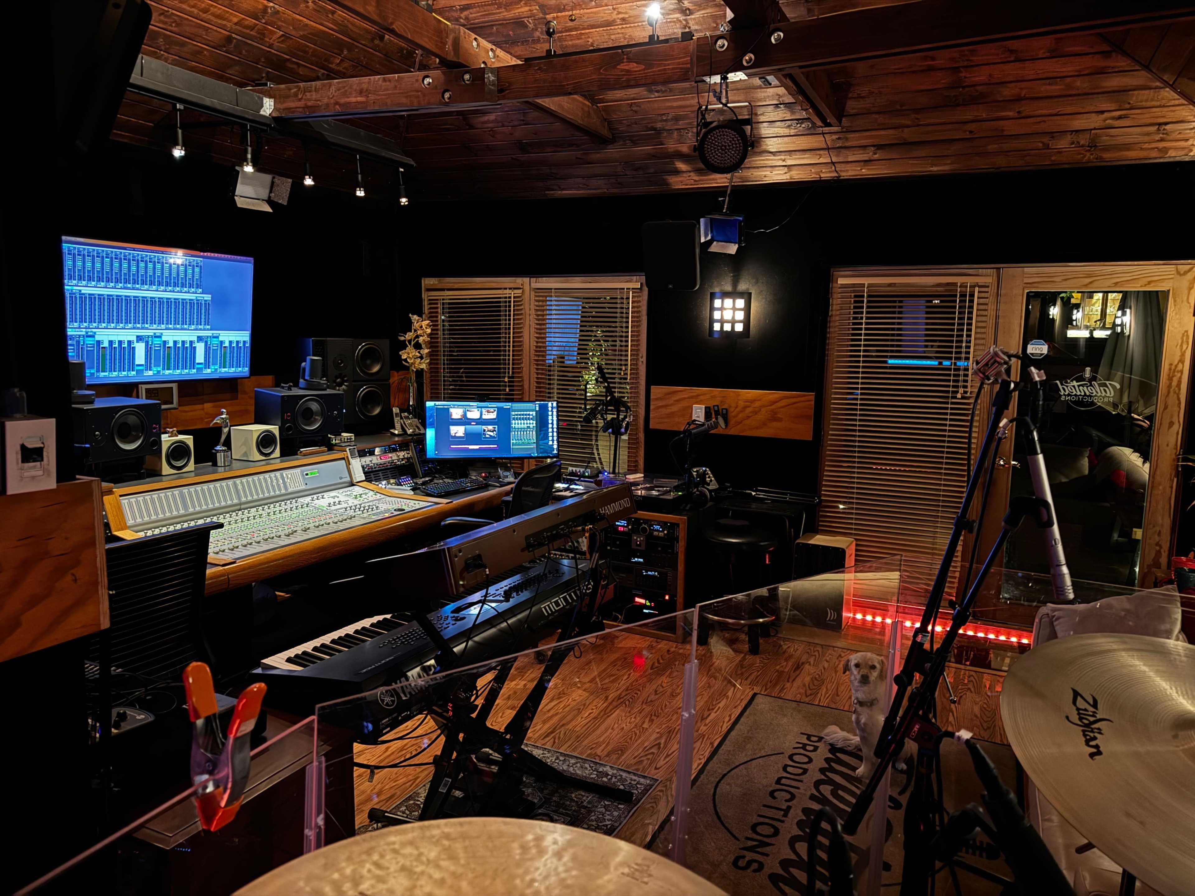 The image shows a well-equipped music recording studio featuring a large mixing console, multiple speakers, and a computer setup, with drums and a keyboard positioned in the foreground.