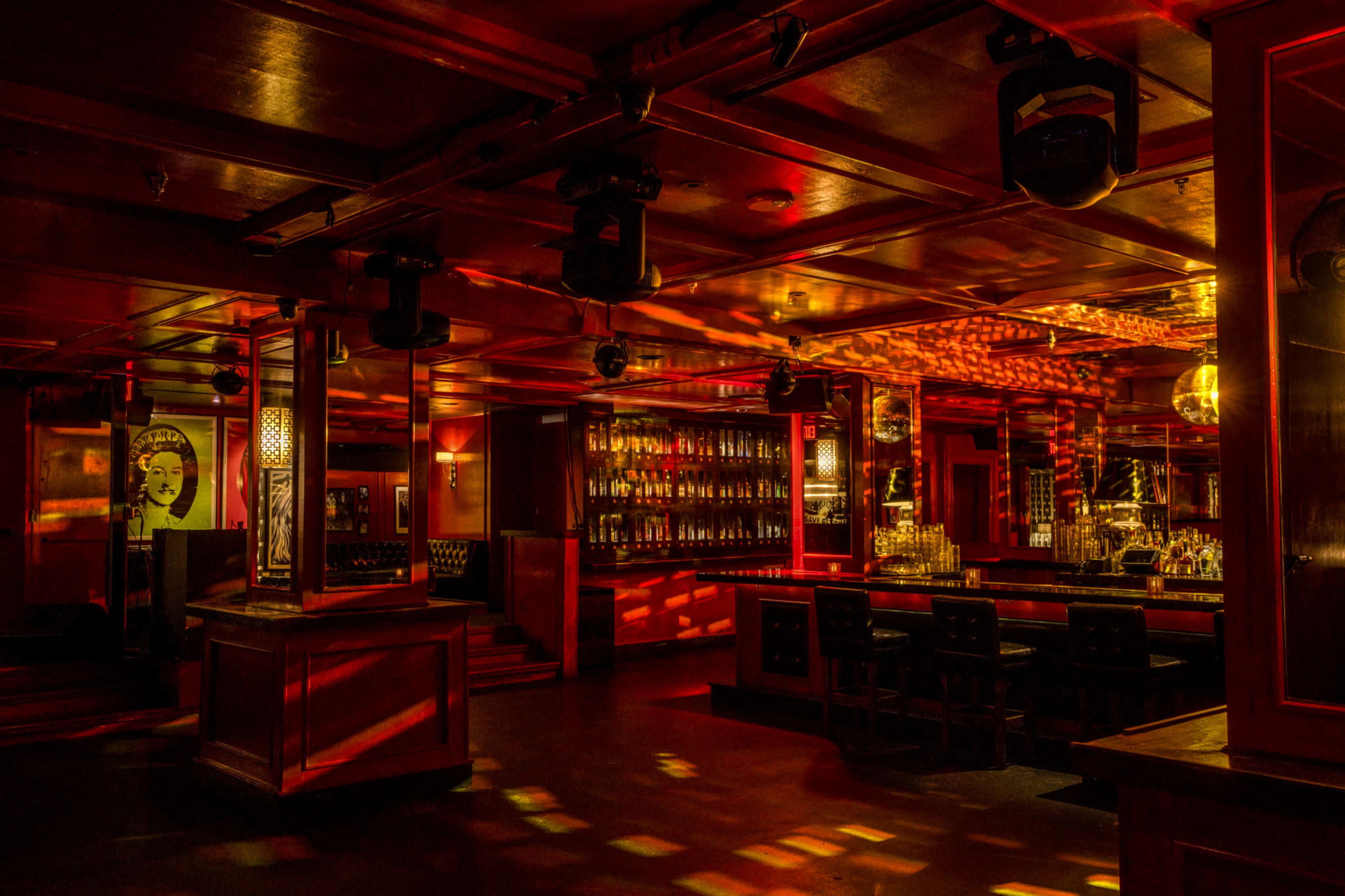 The interior of a dimly lit bar features a long counter lined with bottles, illuminated by warm red lighting and reflected patterns on the walls.