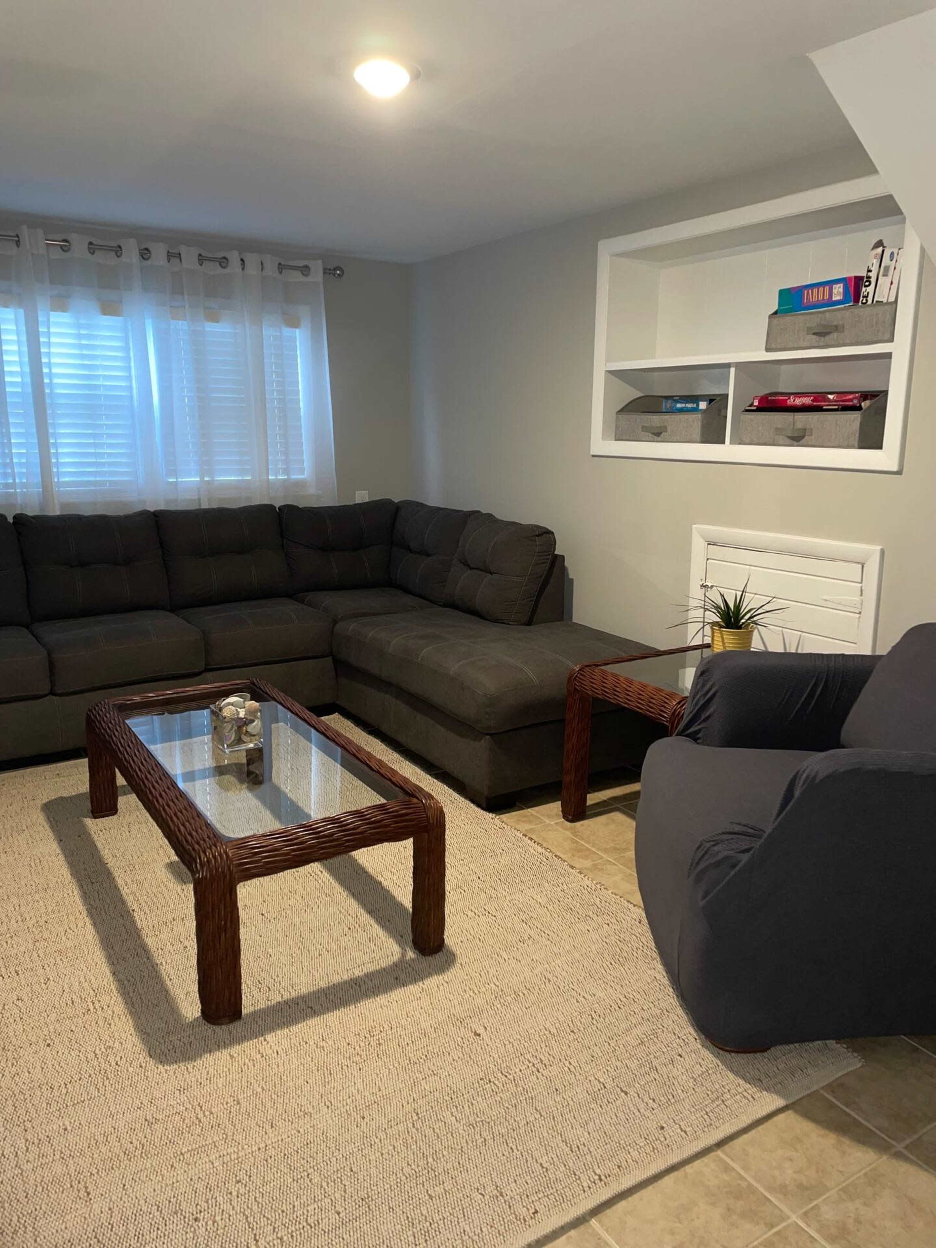 A living room with a sectional sofa, a glass coffee table, and two chairs, complemented by a light-colored area rug and shelves on the wall.