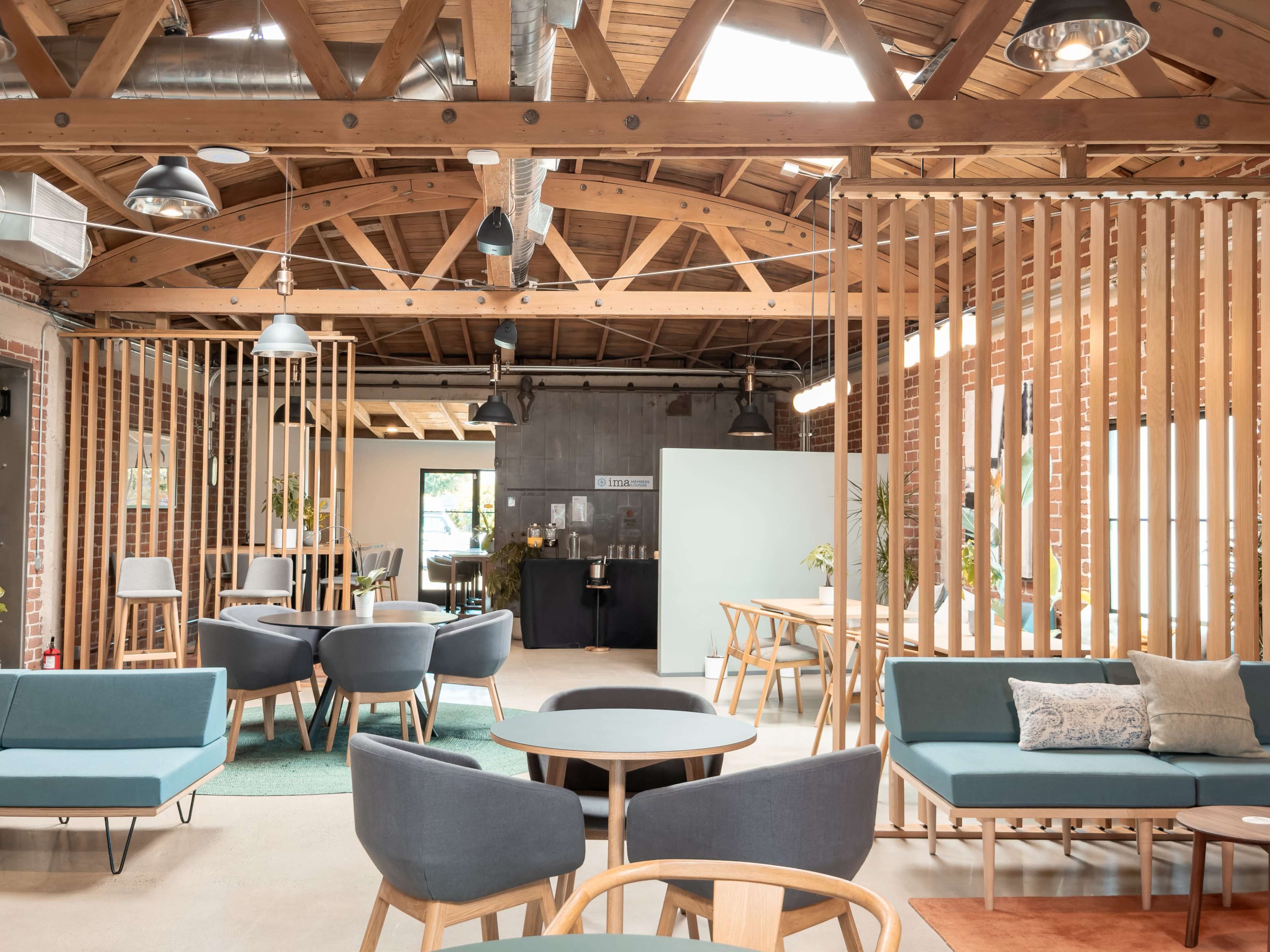 The image features a modern, open-plan workspace with wooden beams, multiple seating areas, and a kitchen area in the background.