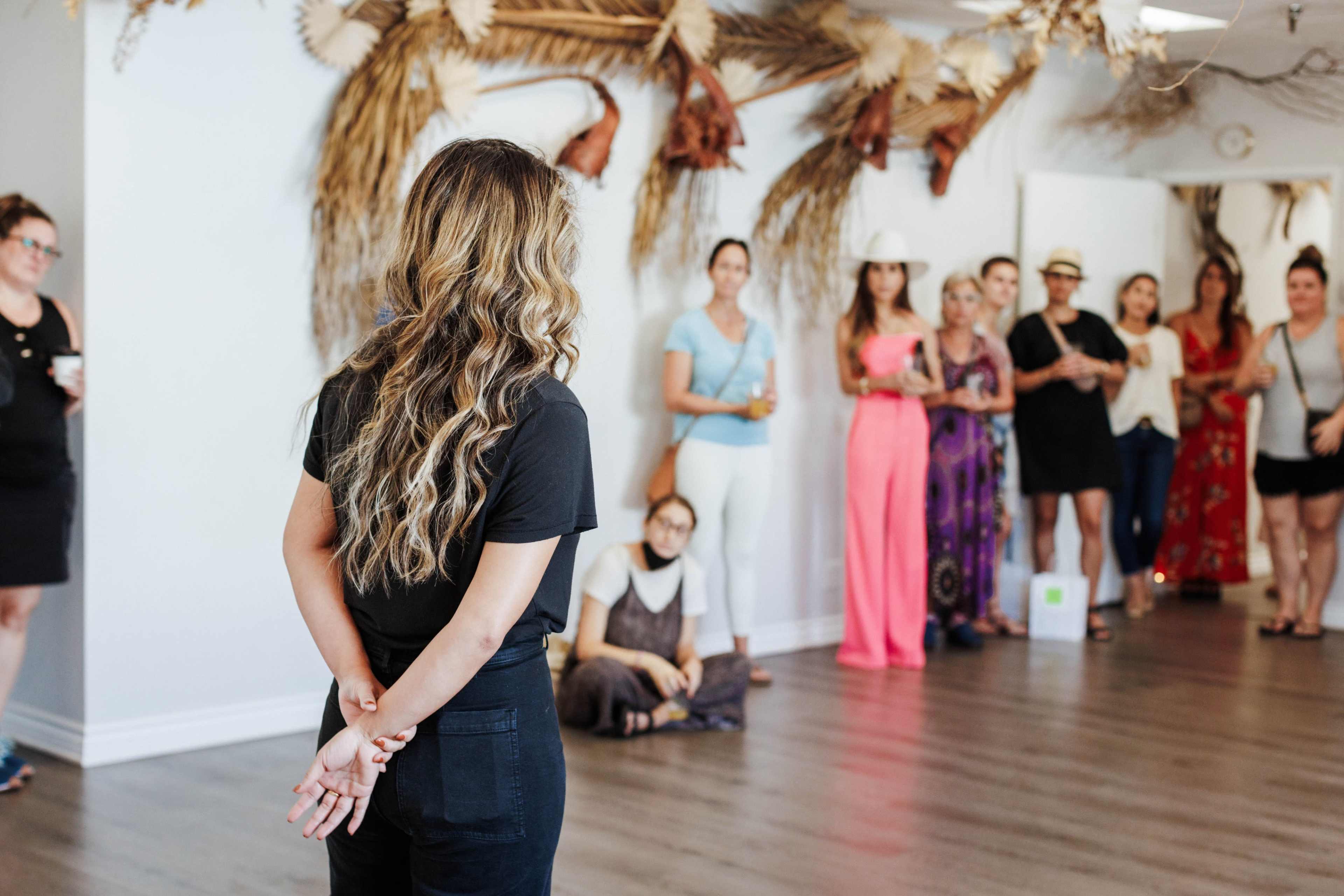 A woman with long hair stands facing a group of people in a decorated room with plants and artistic elements on the walls.