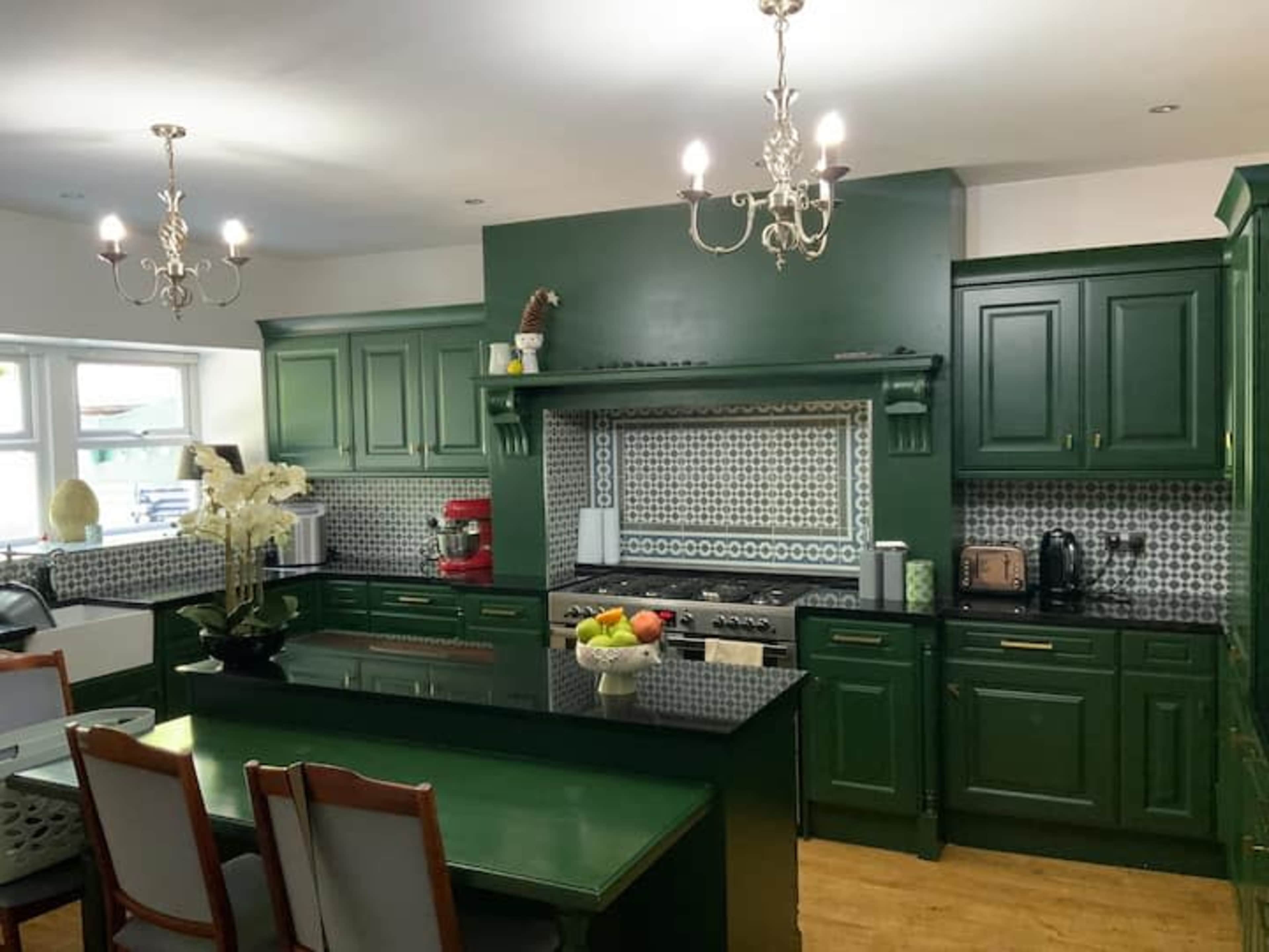 The kitchen features green cabinetry, a black countertop, and decorative tile backsplashes with two chandeliers overhead.