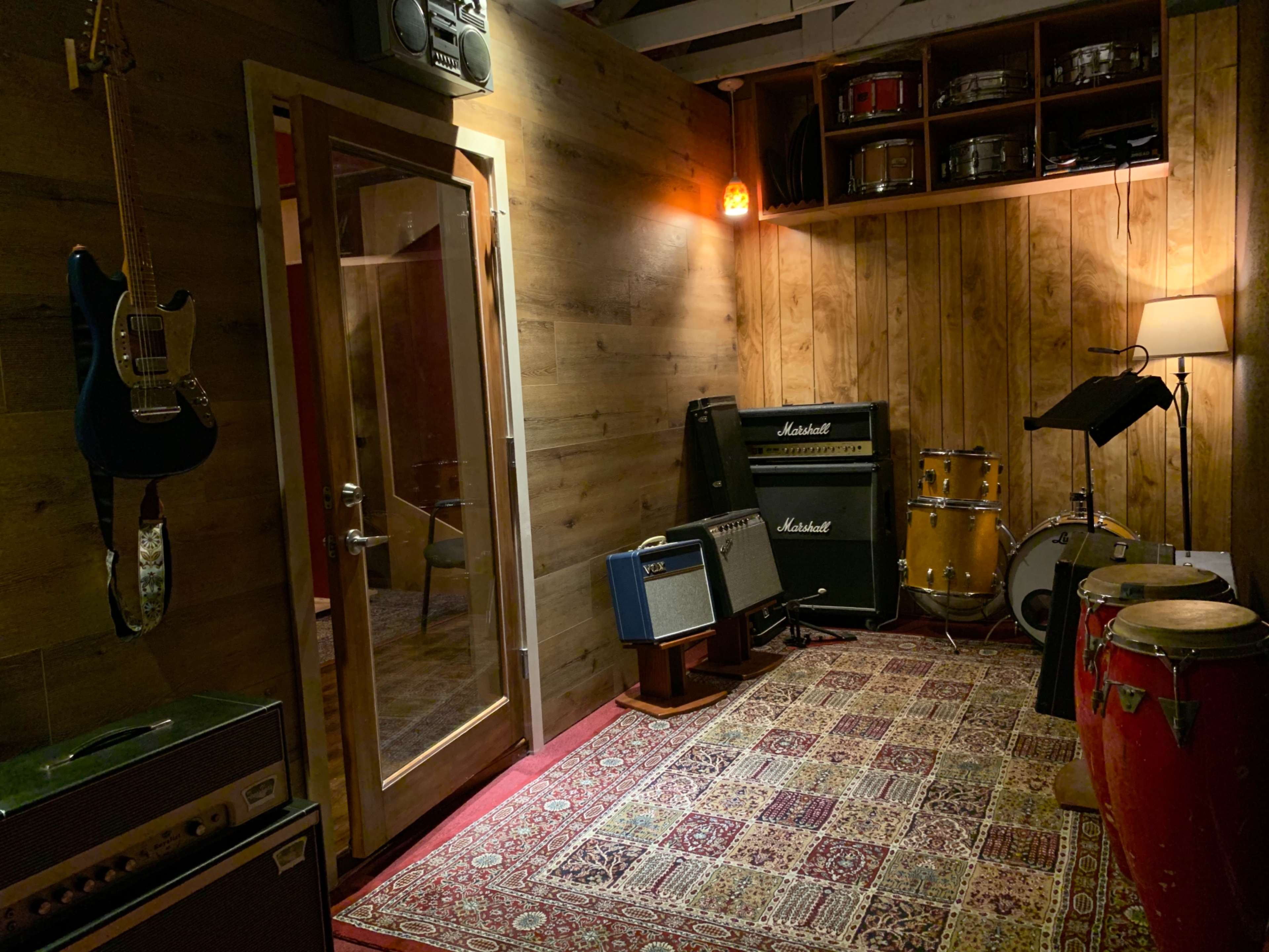 A music rehearsal room features various instruments, including electric guitars, amplifiers, and a drum set, in a wood-paneled space with a patterned rug.
