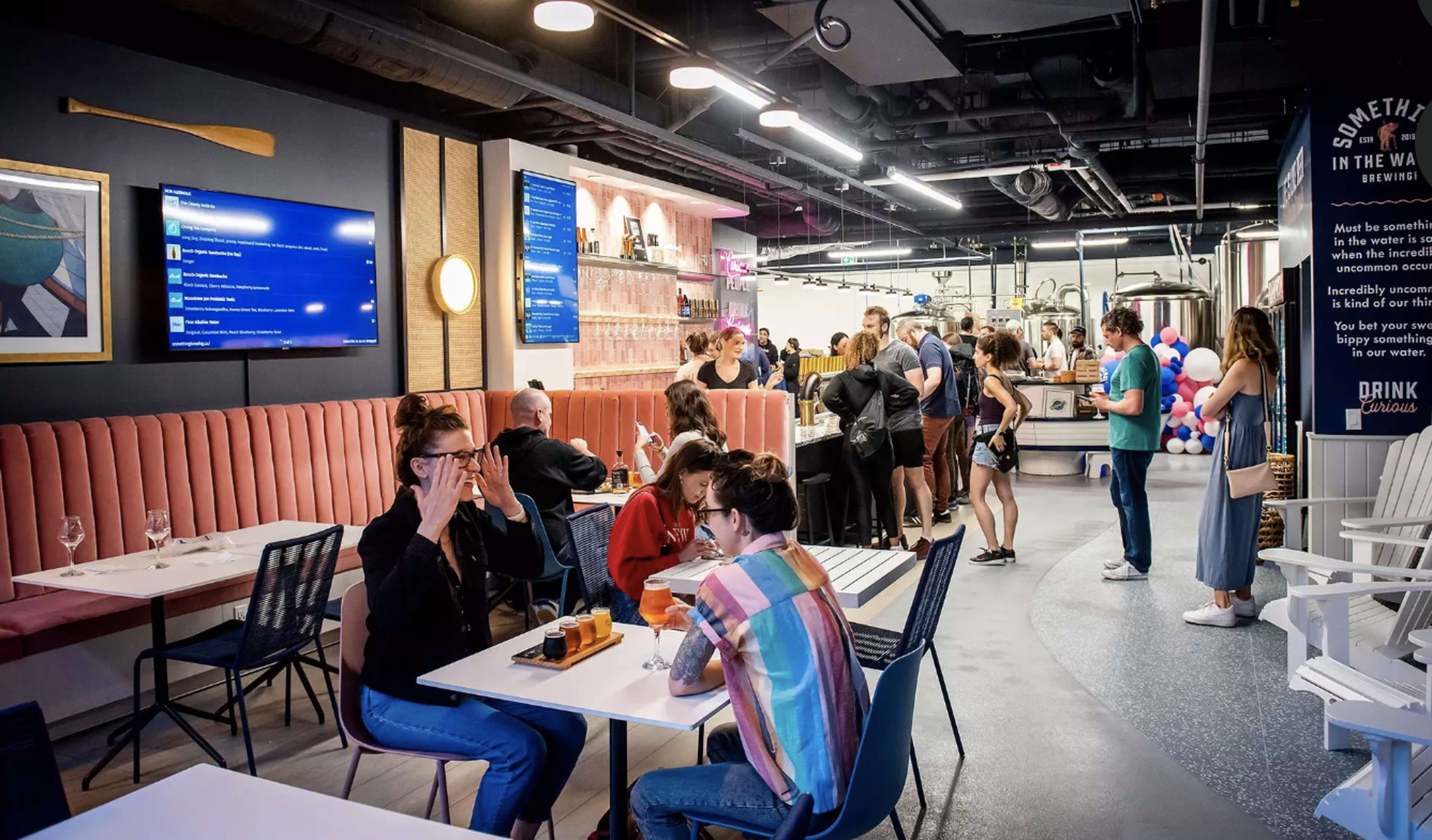 A busy modern bar features a mix of patrons socializing at tables and a long counter with staff serving drinks.