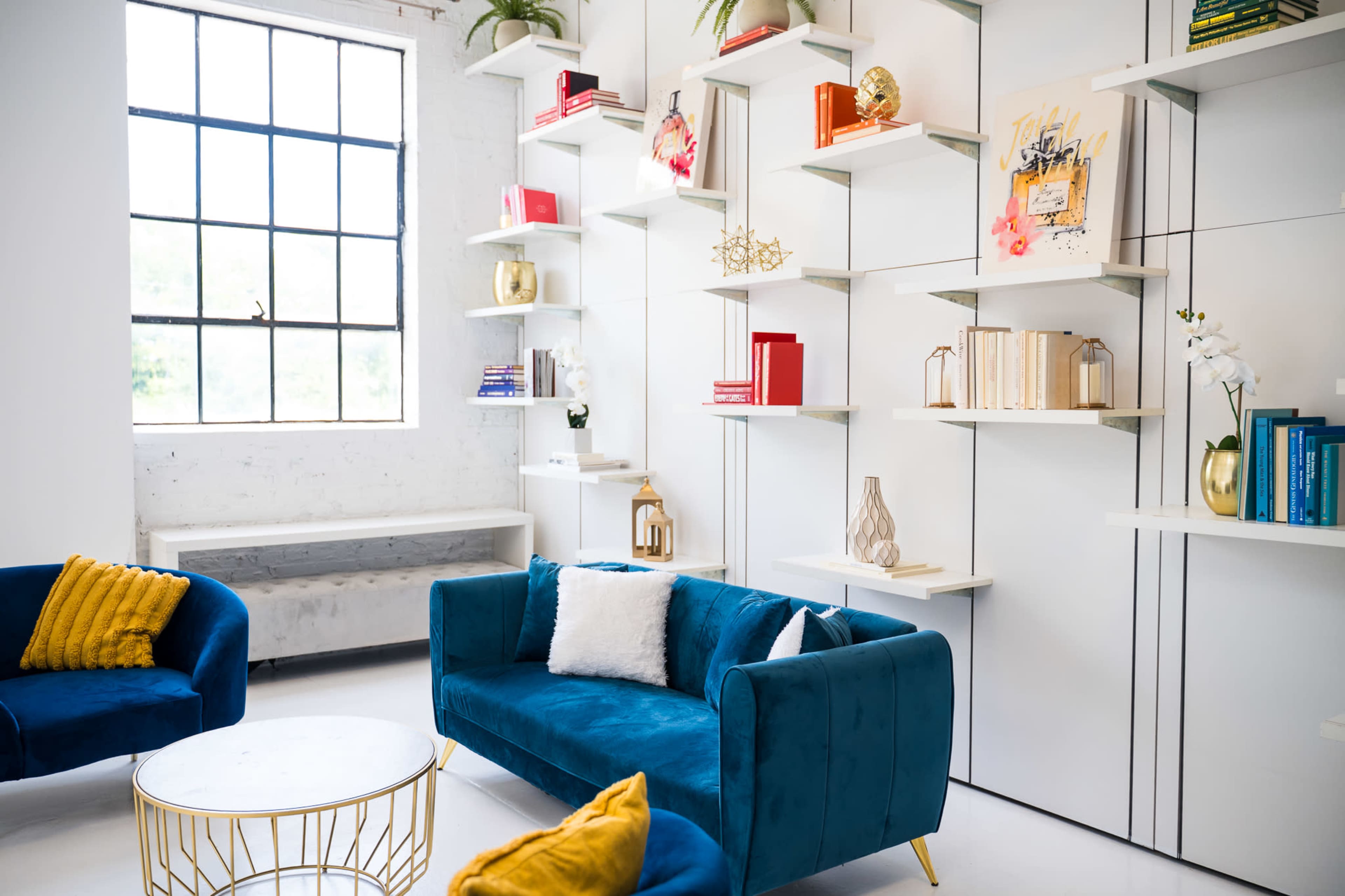 The image shows a modern interior living space featuring a bright window, a stylish blue sofa, two blue chairs, and a decorative shelving unit displaying books and decorative items.