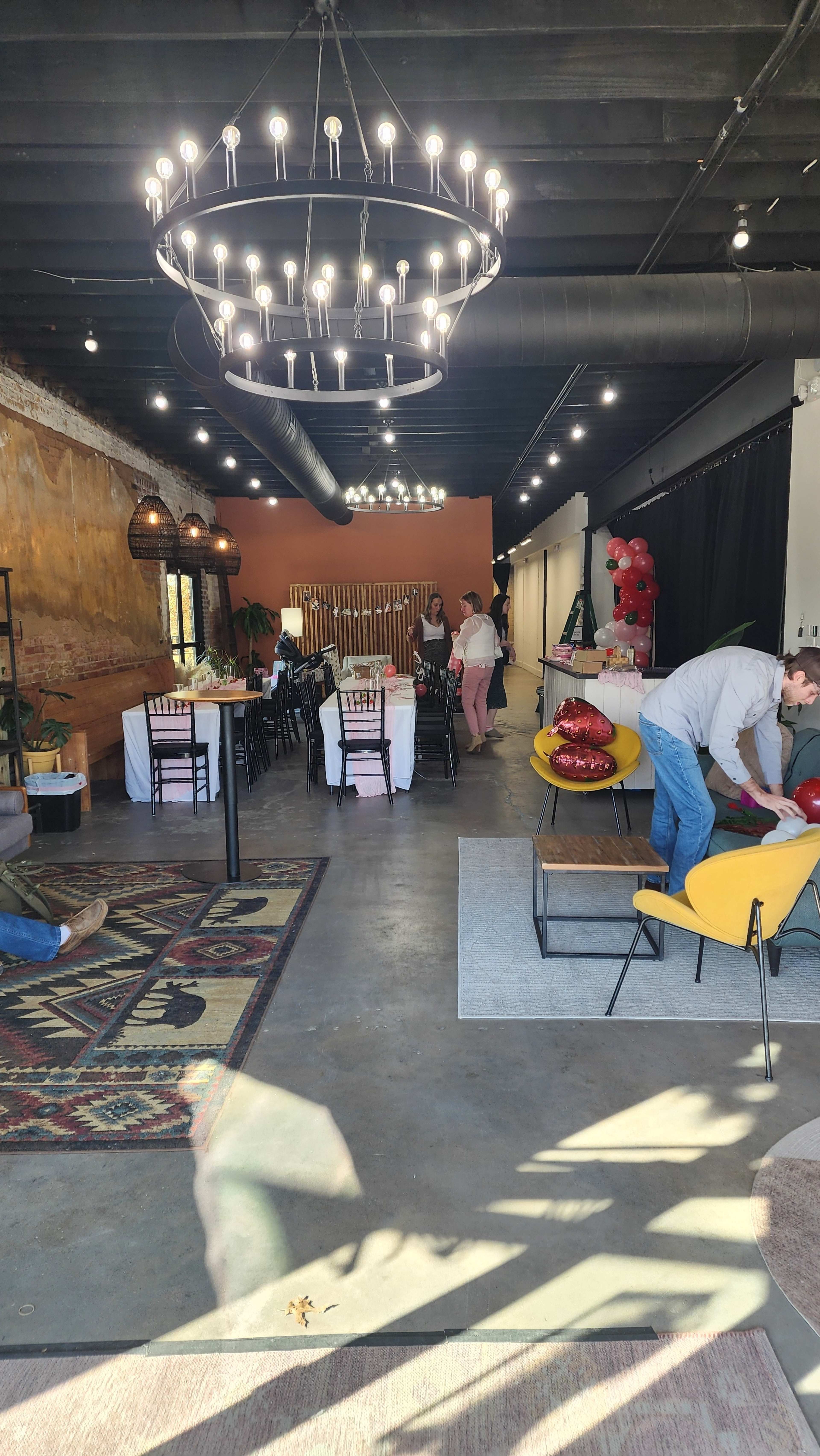 The interior of an event space features a dining area with tables set for a gathering, pendant lights hanging from the ceiling, and people preparing decorations and seating.