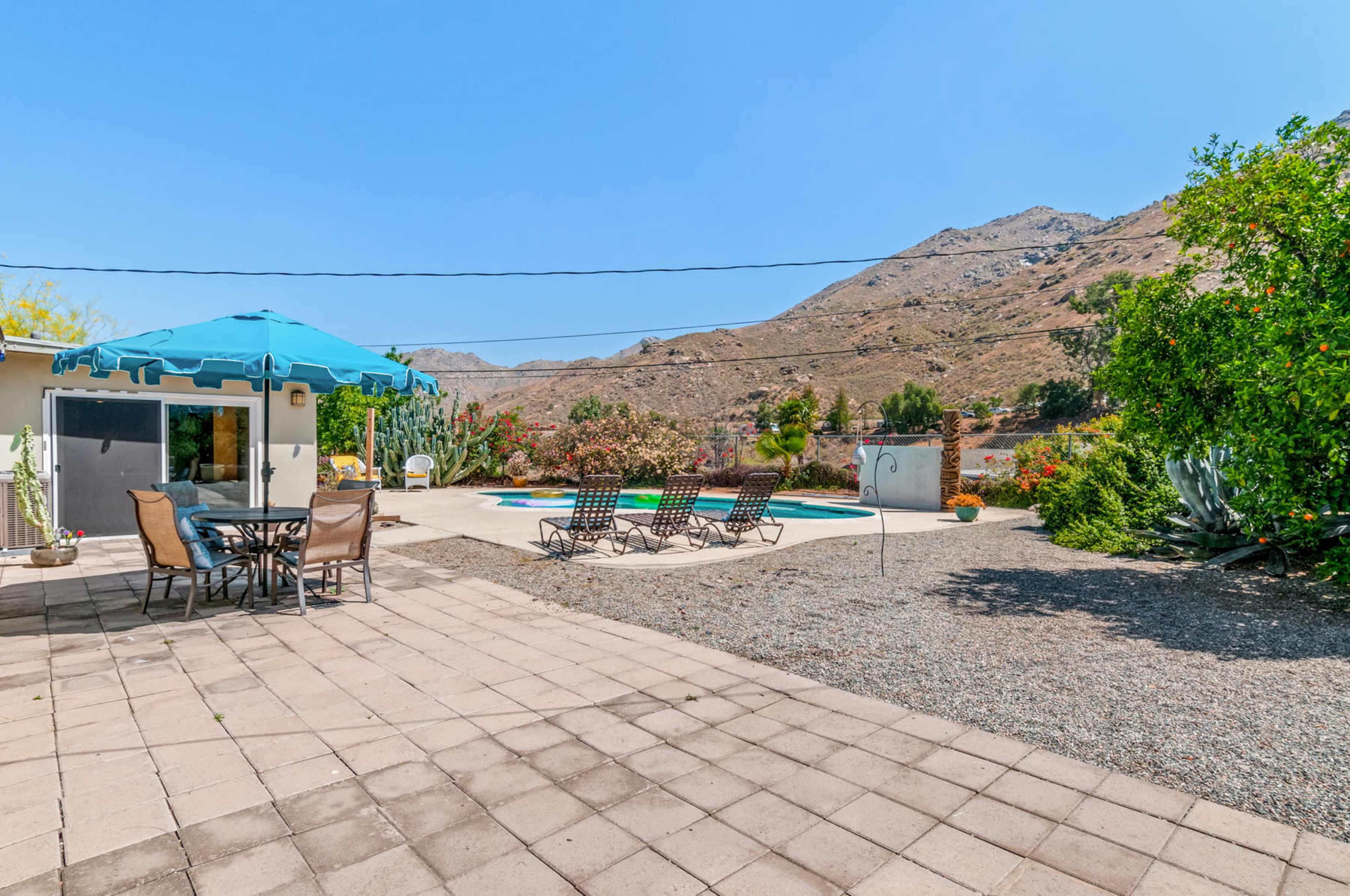 A swimming pool surrounded by lounge chairs is set against a mountainous backdrop with a patio area featuring a table and umbrella.