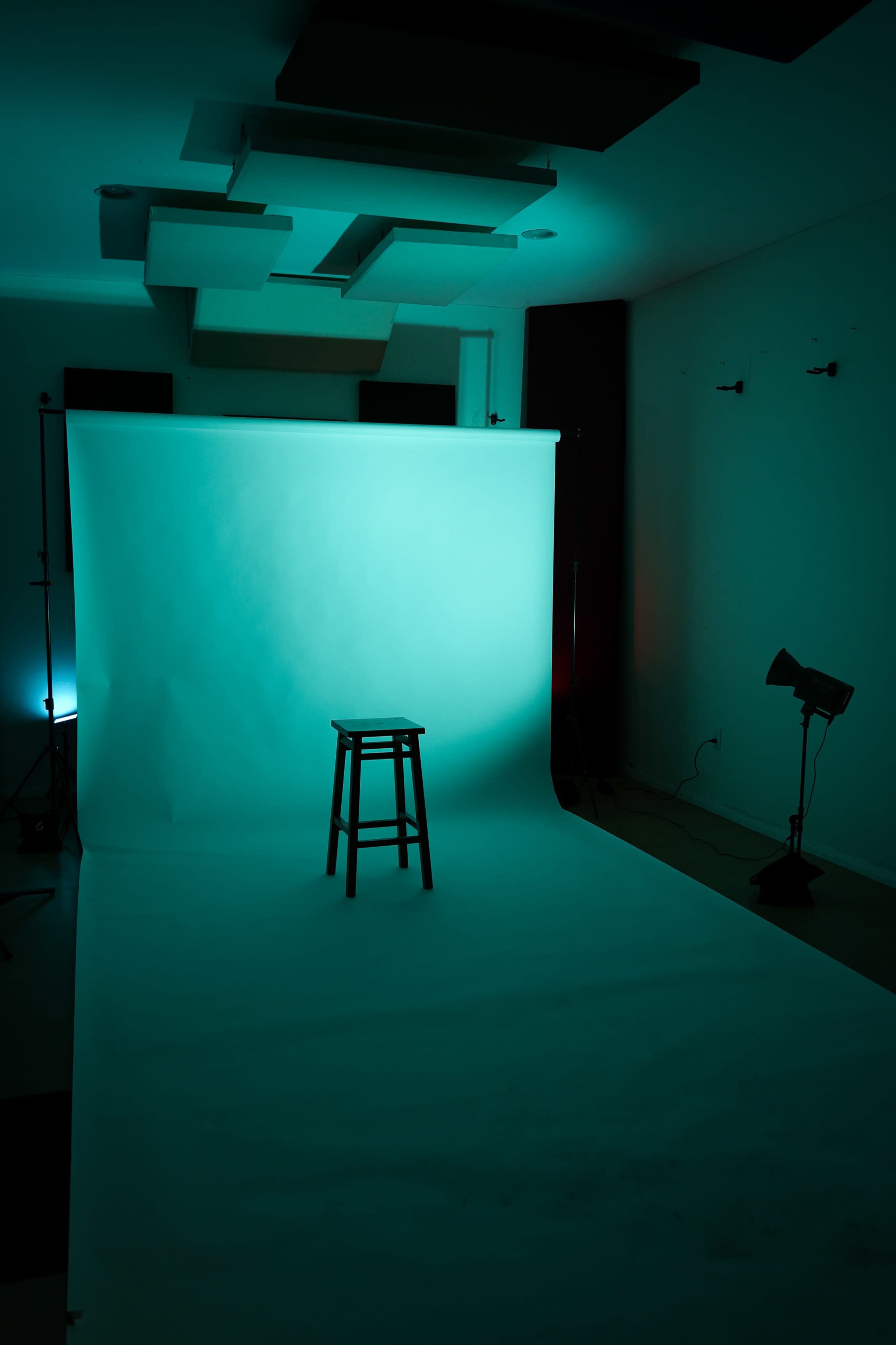 The image shows a minimalist photo studio setup with a single wooden stool positioned in front of a turquoise backdrop.