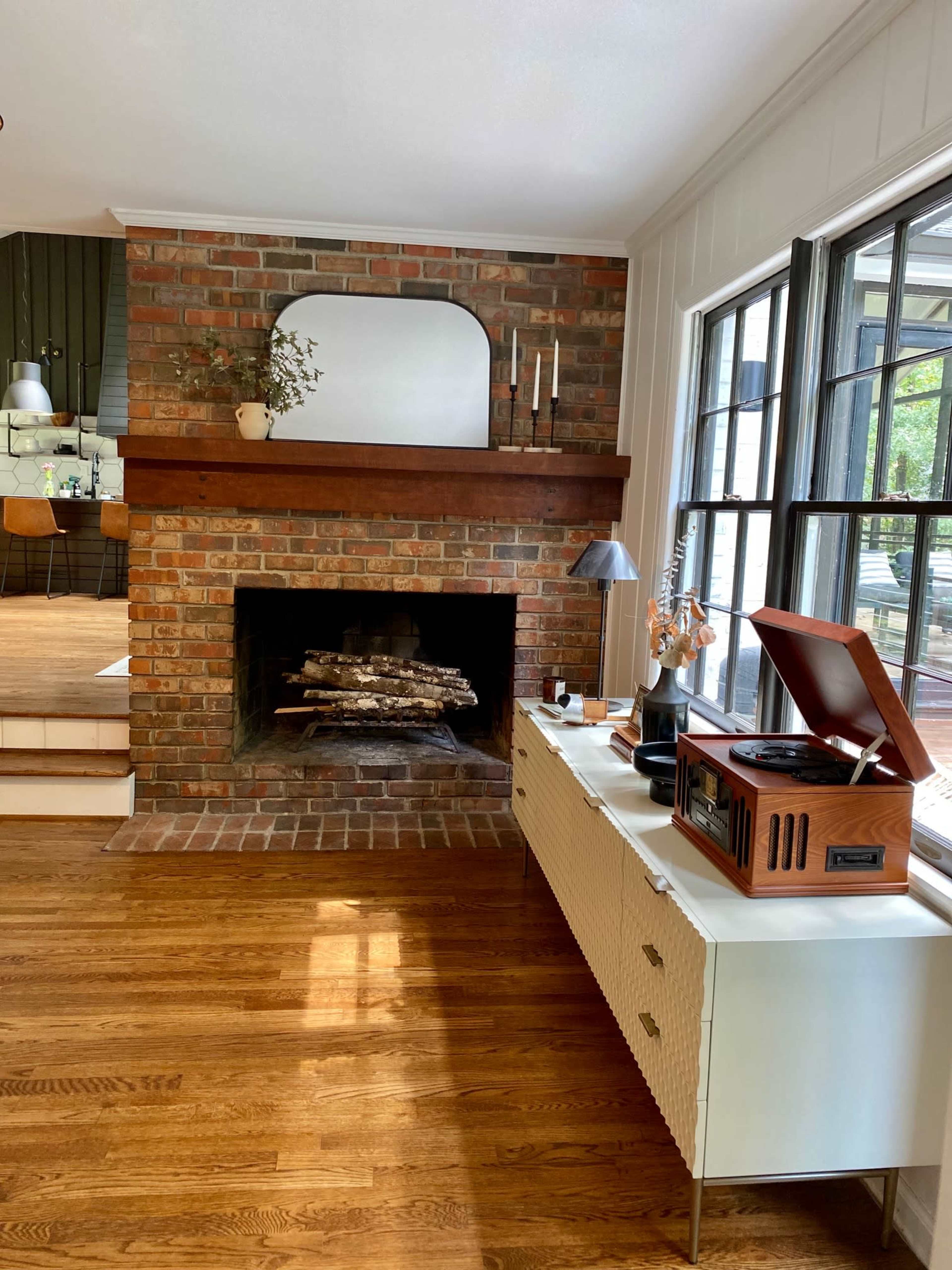 The image shows a living room with a brick fireplace, a wooden mantel, a record player on a white media console, and large windows that allow natural light.