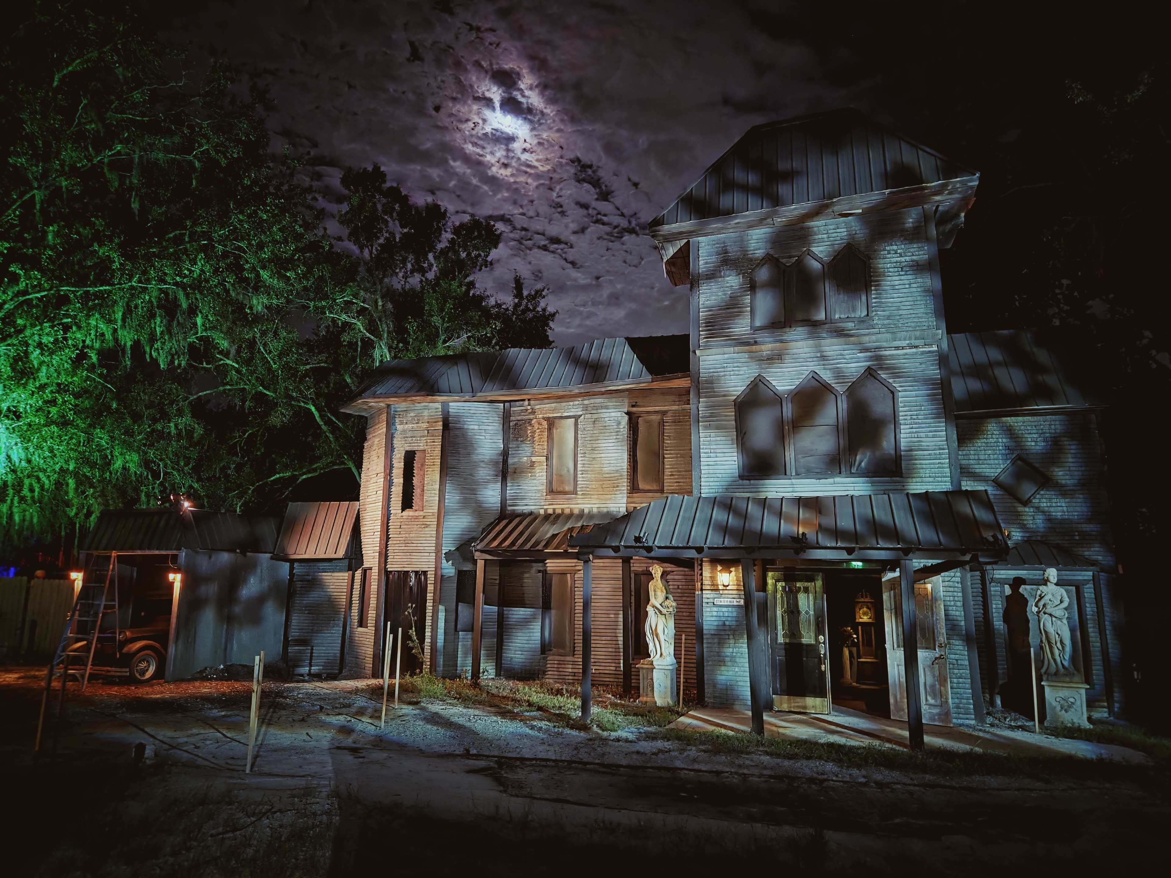 An eerie, two-story brick house with a peaked roof and shadowy figures cast by the moonlight stands under a cloudy night sky, surrounded by trees.