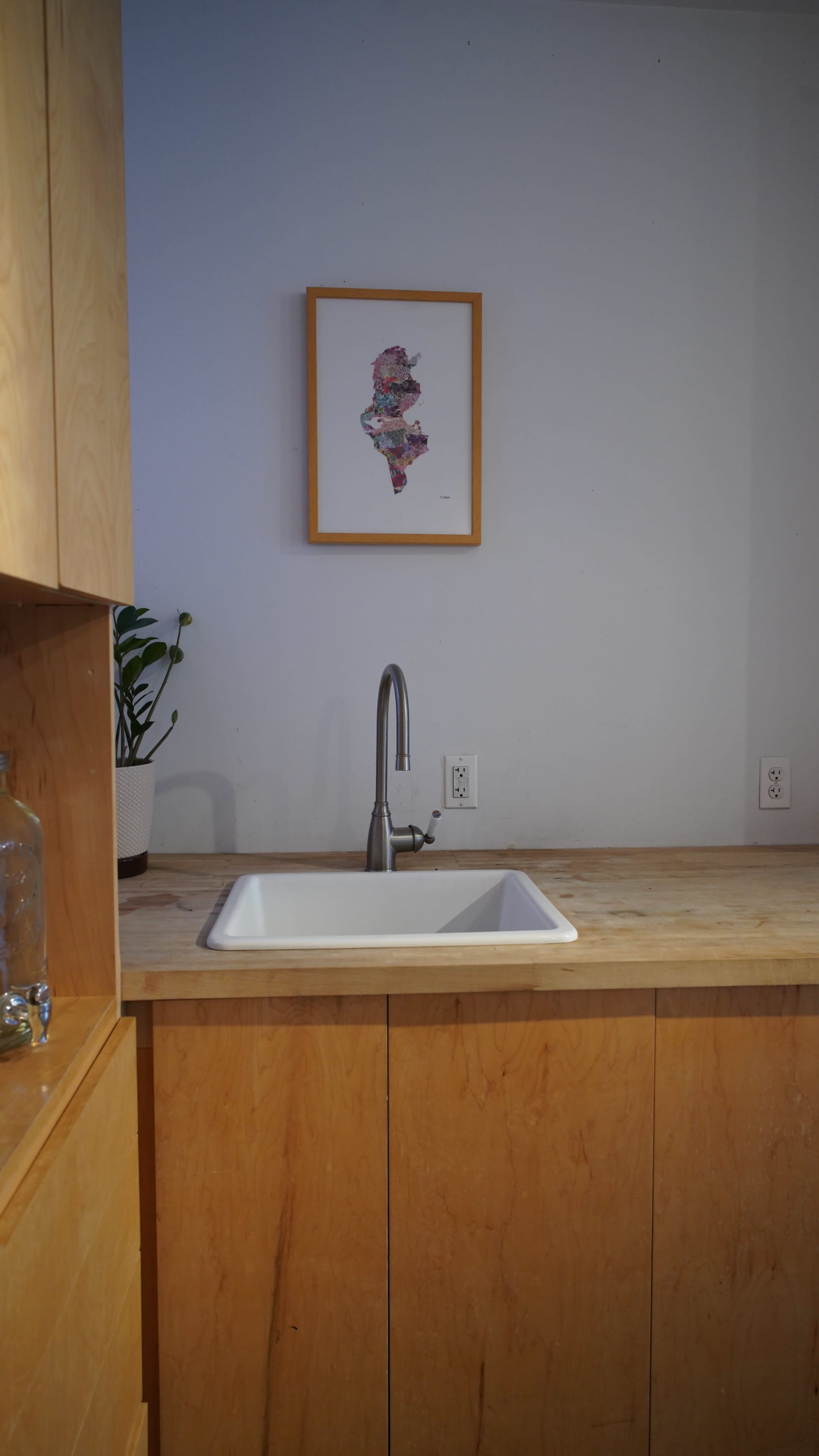The image shows a modern kitchen with a stainless steel sink, a wooden countertop, and a framed artwork on the wall above.