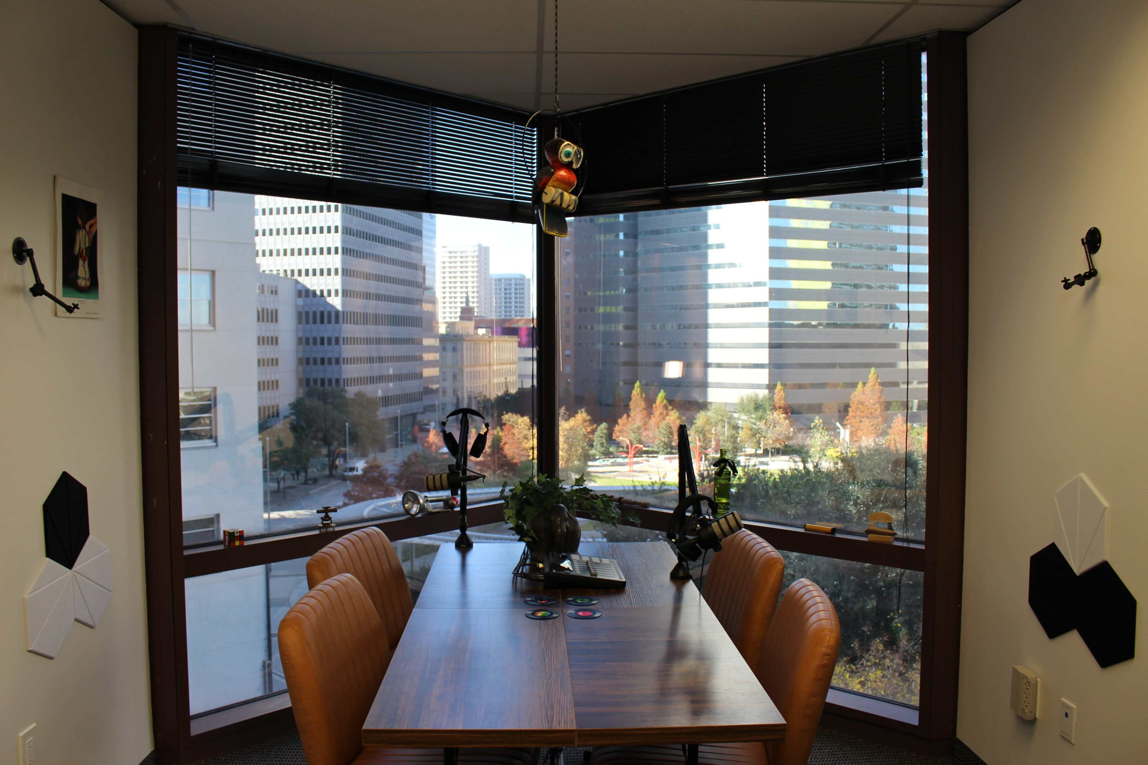 The image shows an office space with a wooden conference table surrounded by leather chairs, large windows offering a view of city buildings and trees outside, and a small potted plant on the table.