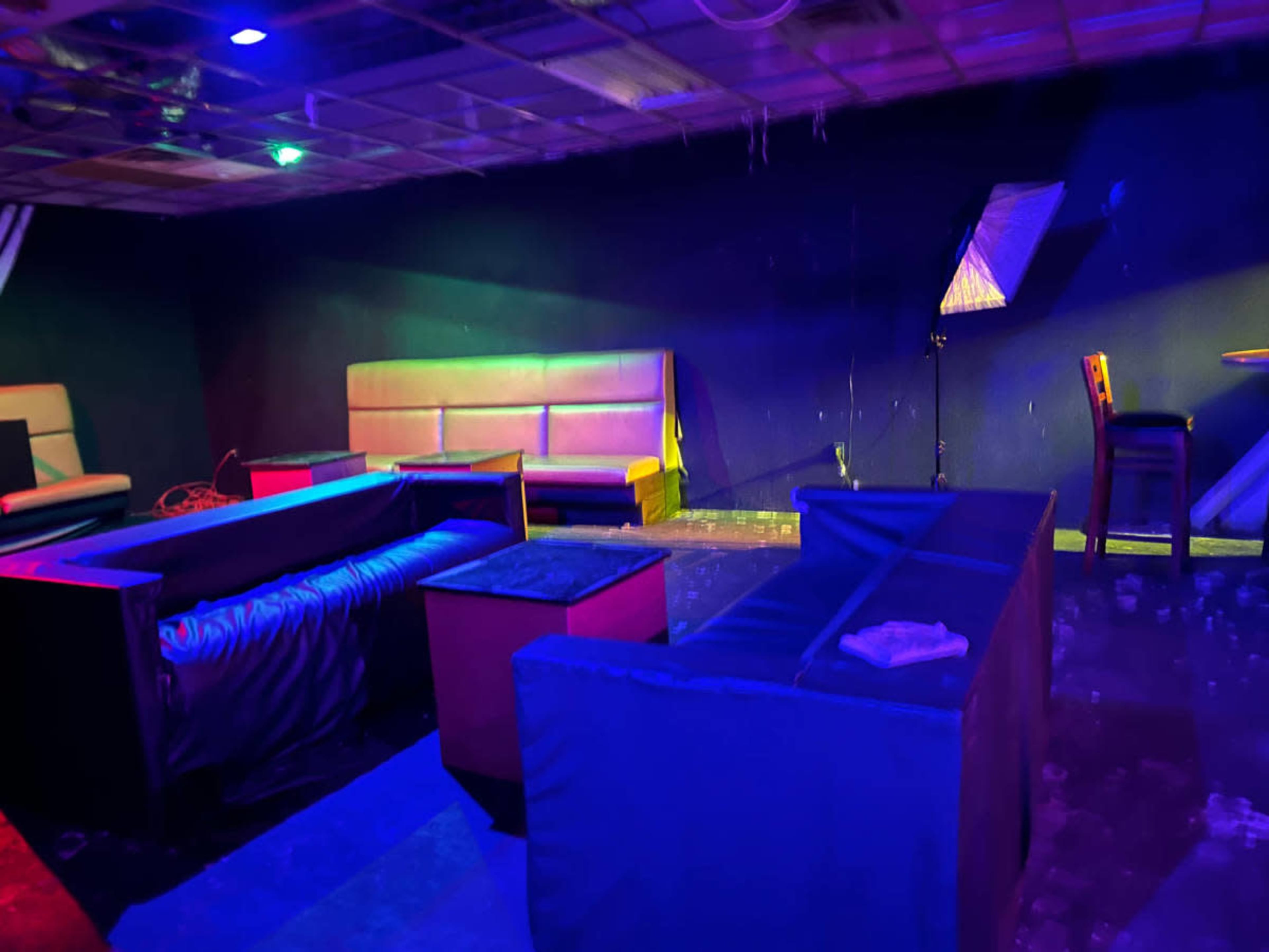 The image shows a dimly lit lounge area with several seating arrangements, including couches and tables, illuminated by colored lights.