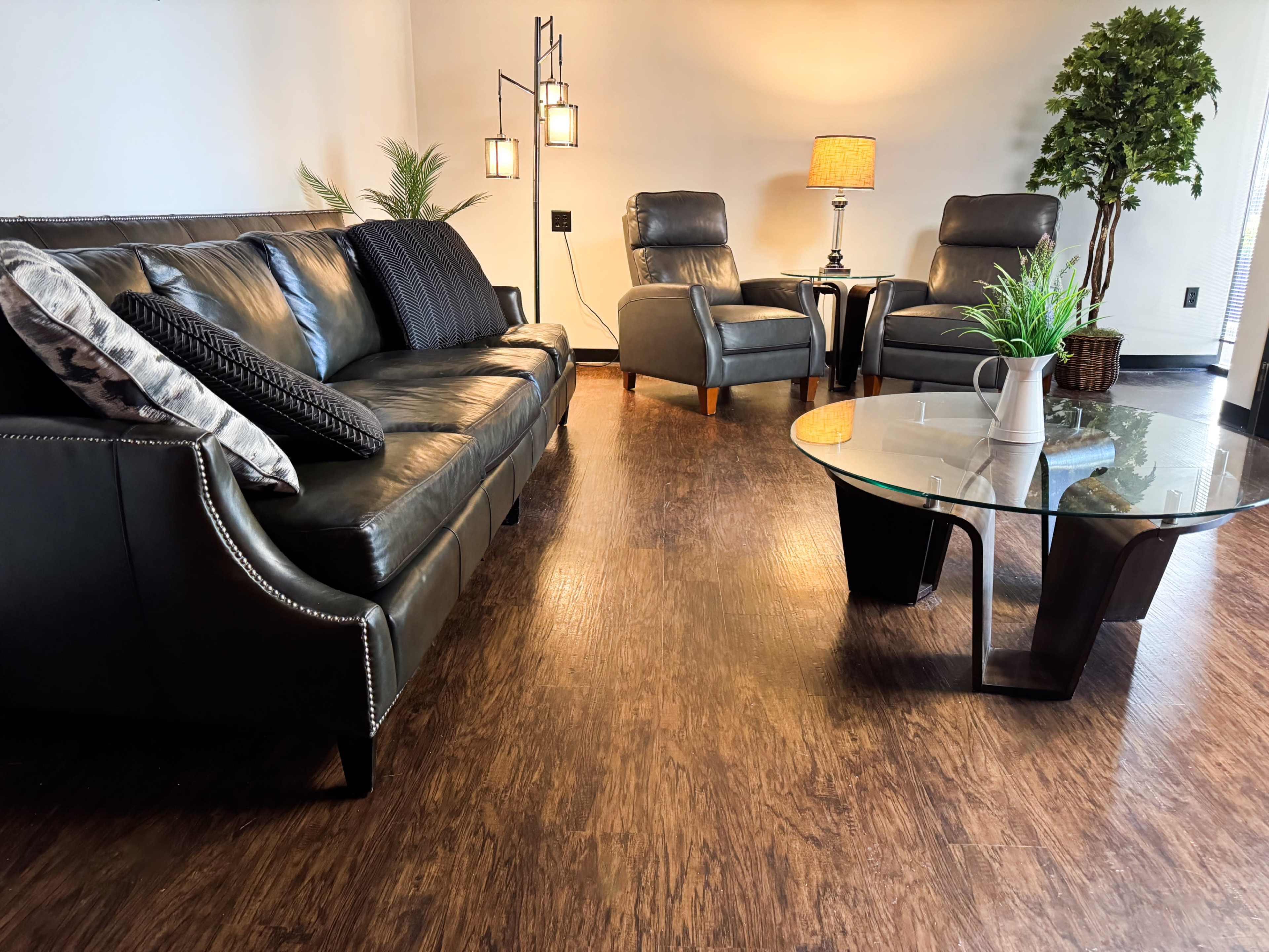 The image shows a modern living room with a black leather sofa, two armchairs, a glass coffee table, and a floor lamp, all set on brown wood-style flooring.