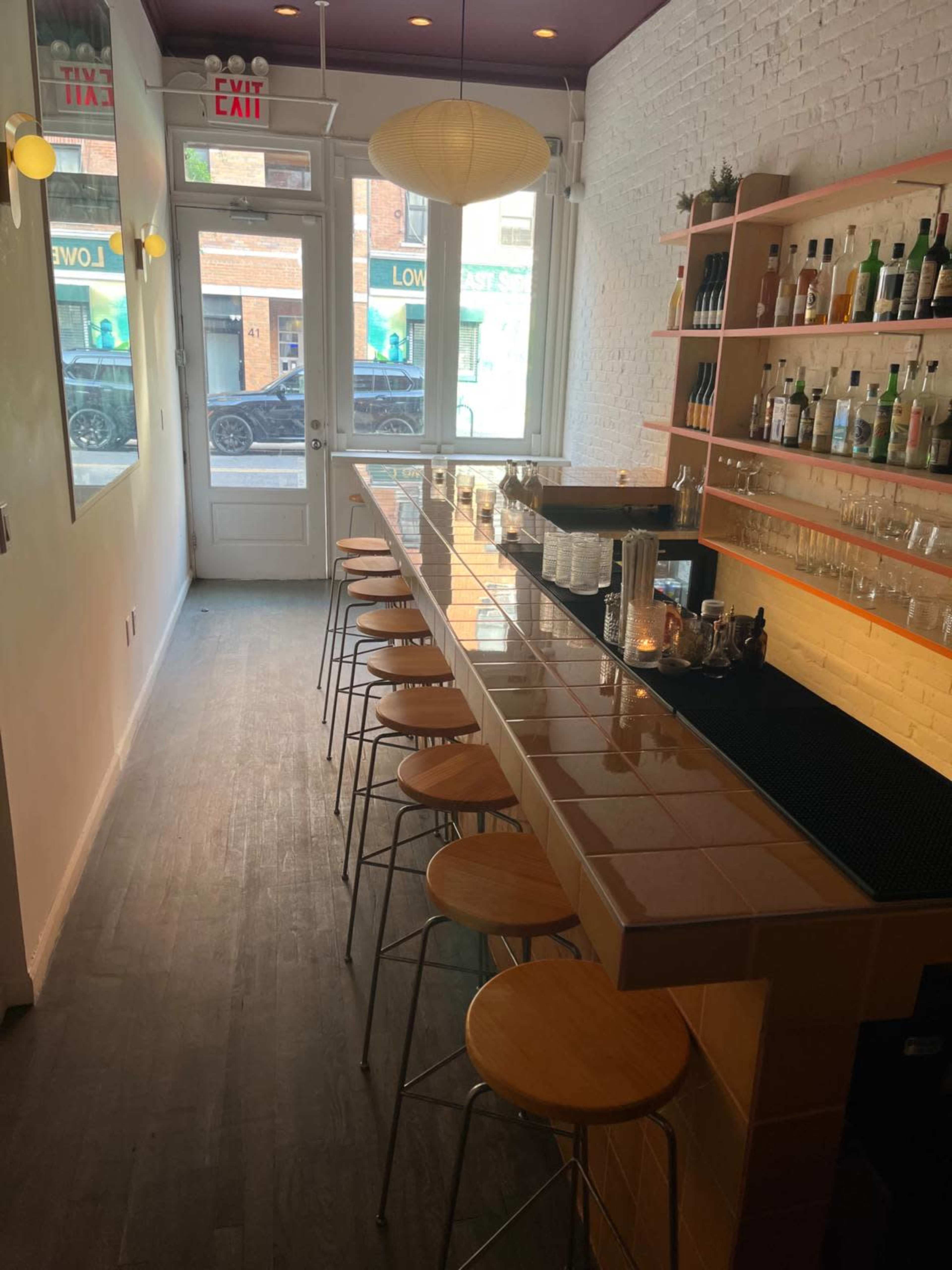 The image shows a narrow, bright bar area with wooden stools, a long counter, and a wall lined with bottles on shelves.