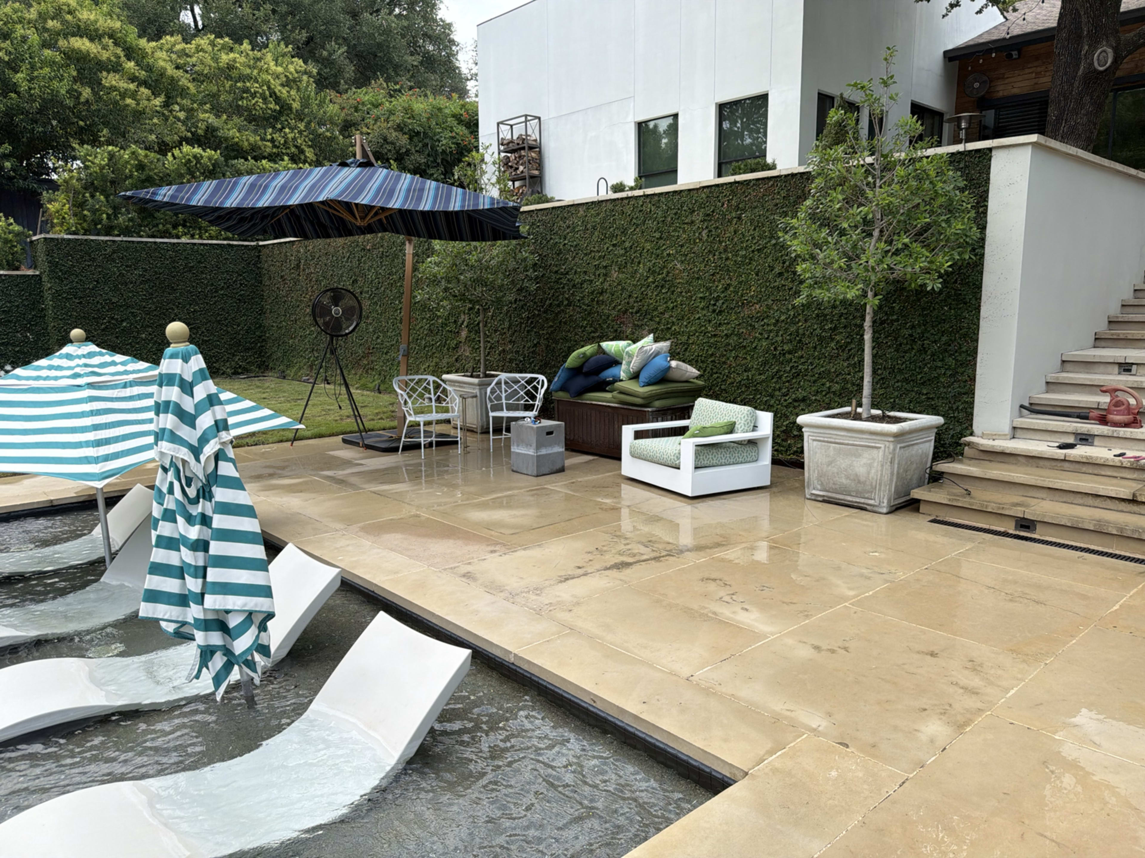 The image shows a patio area with a pool featuring lounge chairs, a table with two white chairs, green cushions on a sofa, an umbrella, and a fan near a hedge and a modern white building.