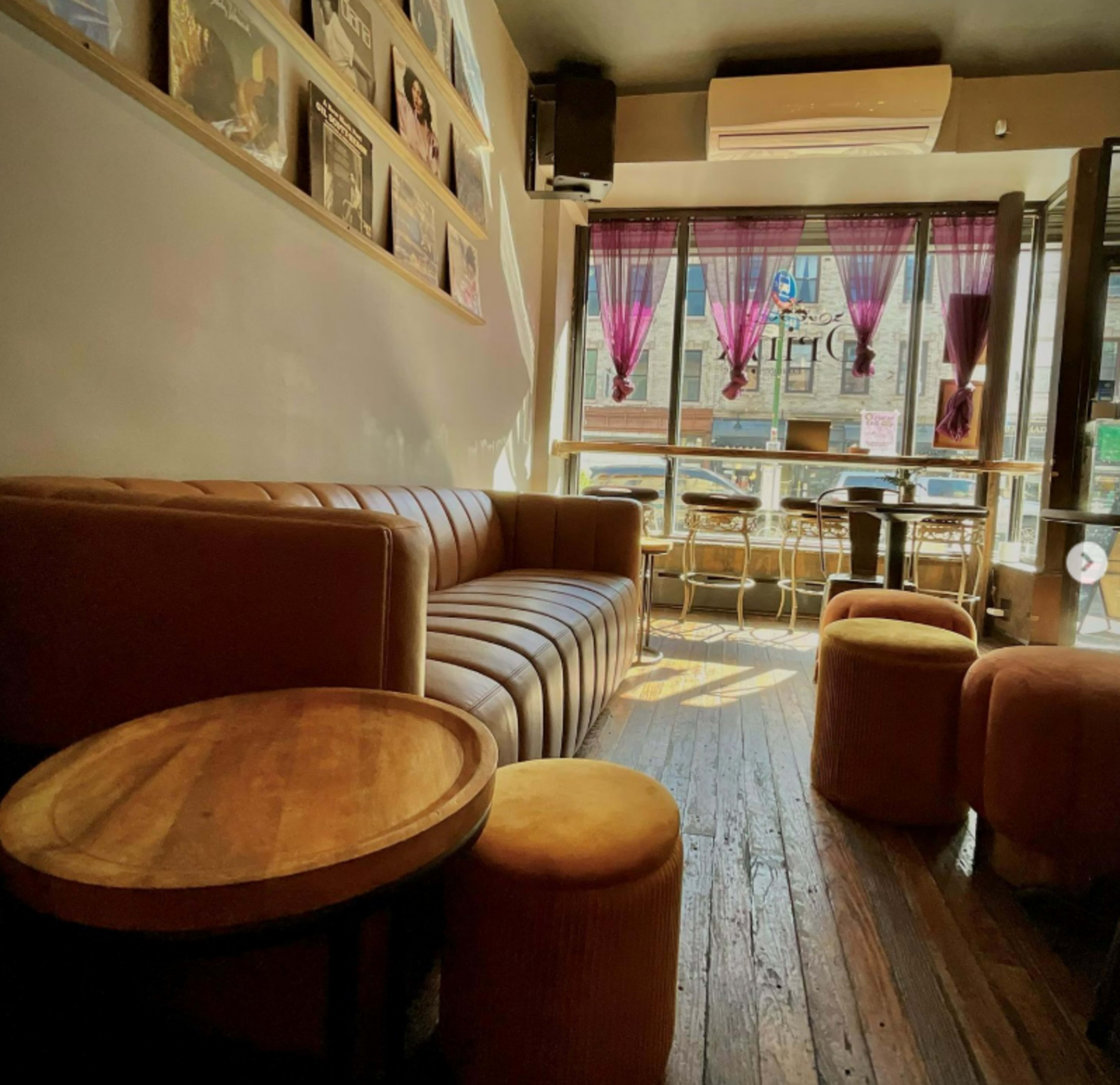 The image shows a cozy café interior with a leather sofa, wooden tables, and round seating stools, illuminated by natural light from large windows.