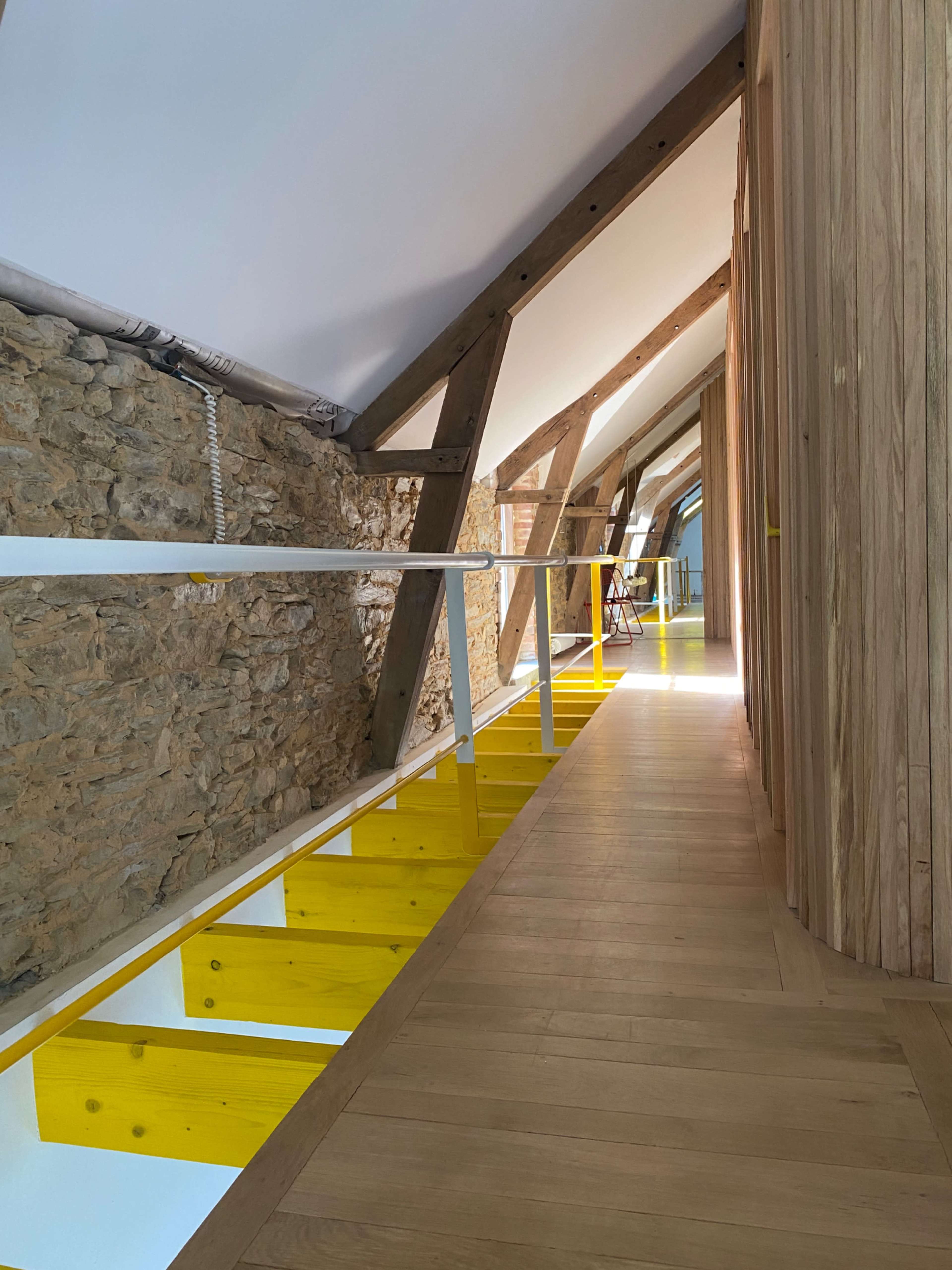 The image shows a hallway with a wooden floor, featuring exposed stone walls and a modern design with yellow beams and railings.
