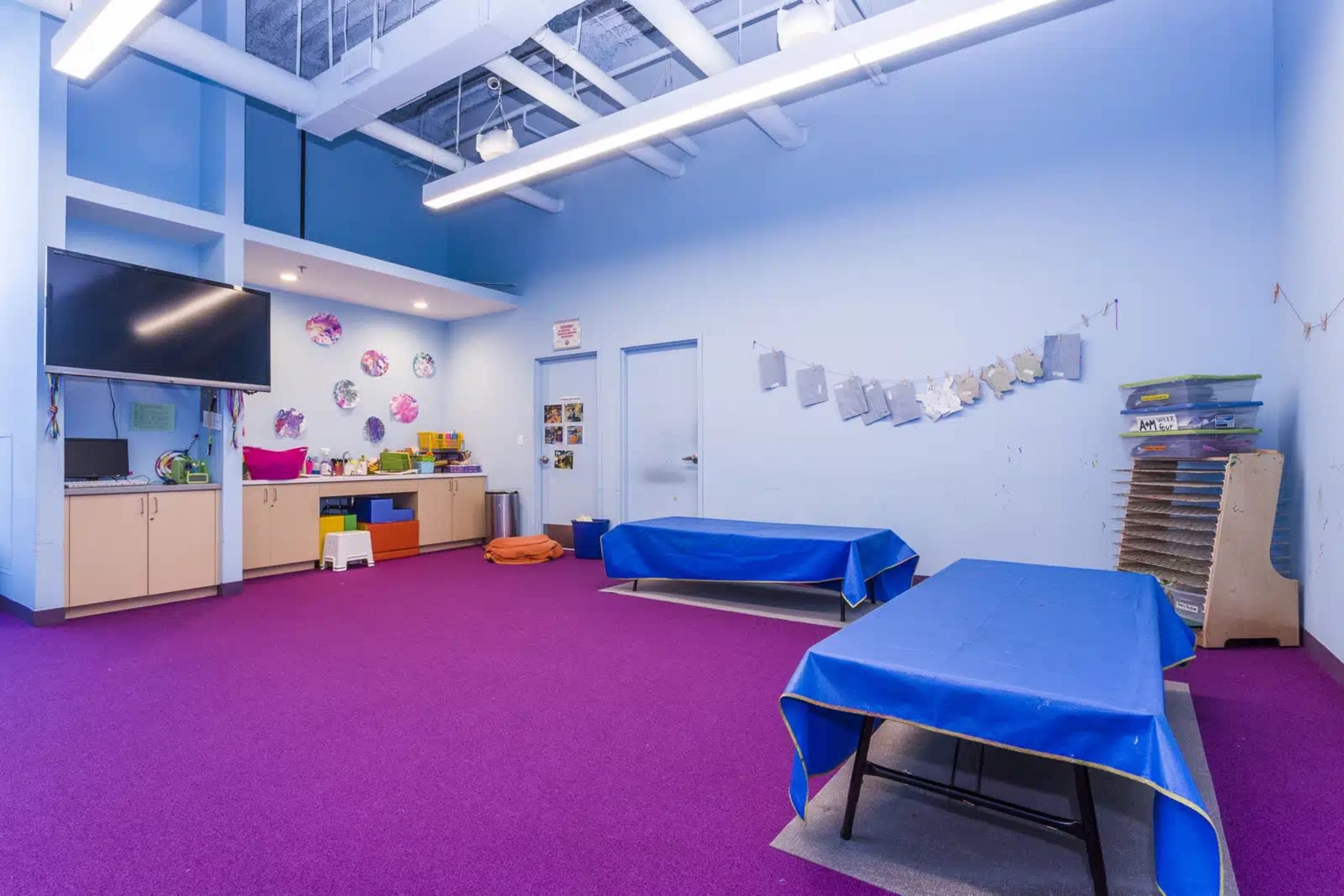 A brightly colored classroom with purple carpet, featuring two blue-covered tables, a television mounted on the wall, and various educational materials scattered around.