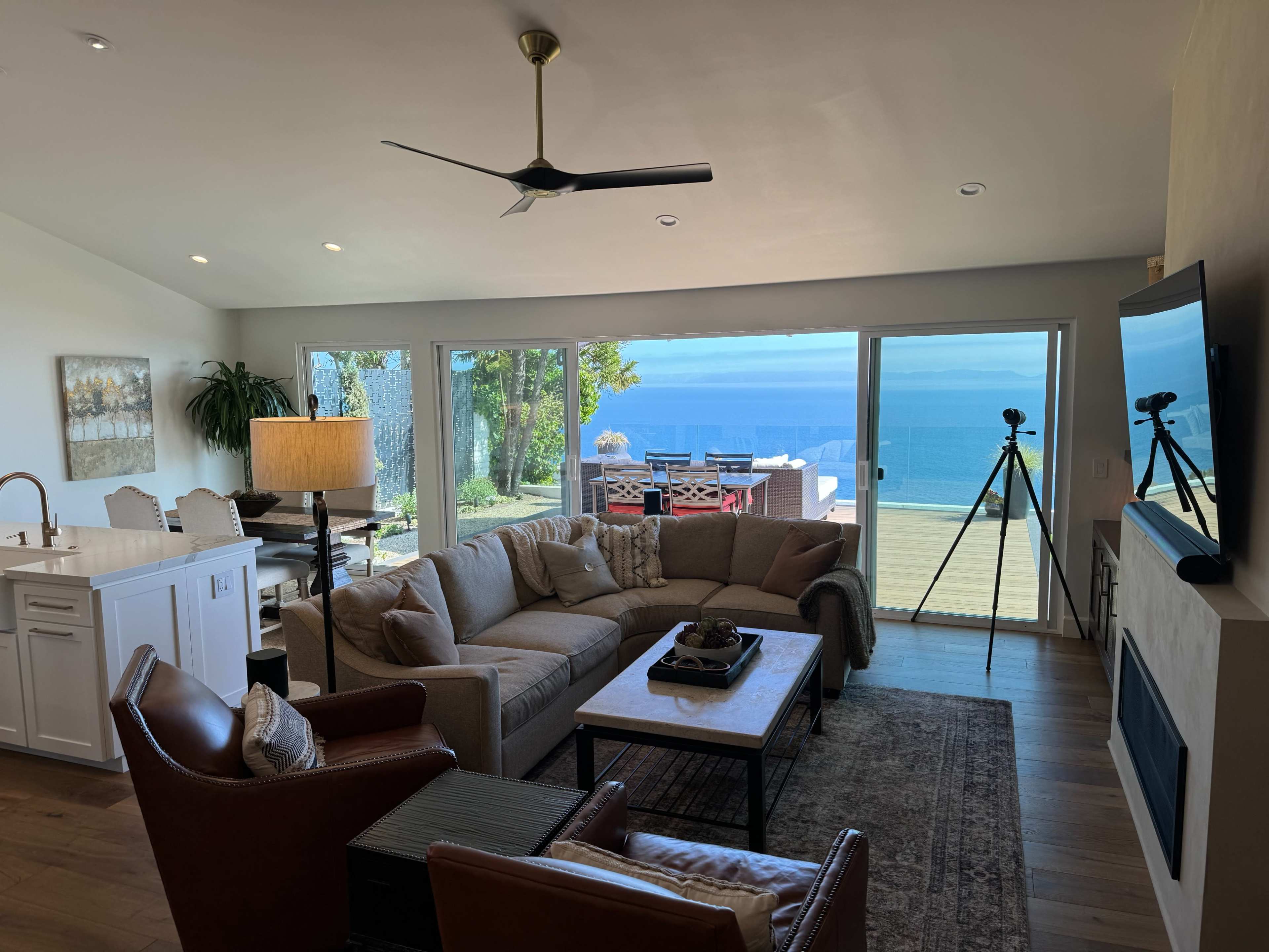 The living area features a large sectional sofa, two armchairs, and a coffee table, with large windows showcasing a view of the ocean and outdoor seating.