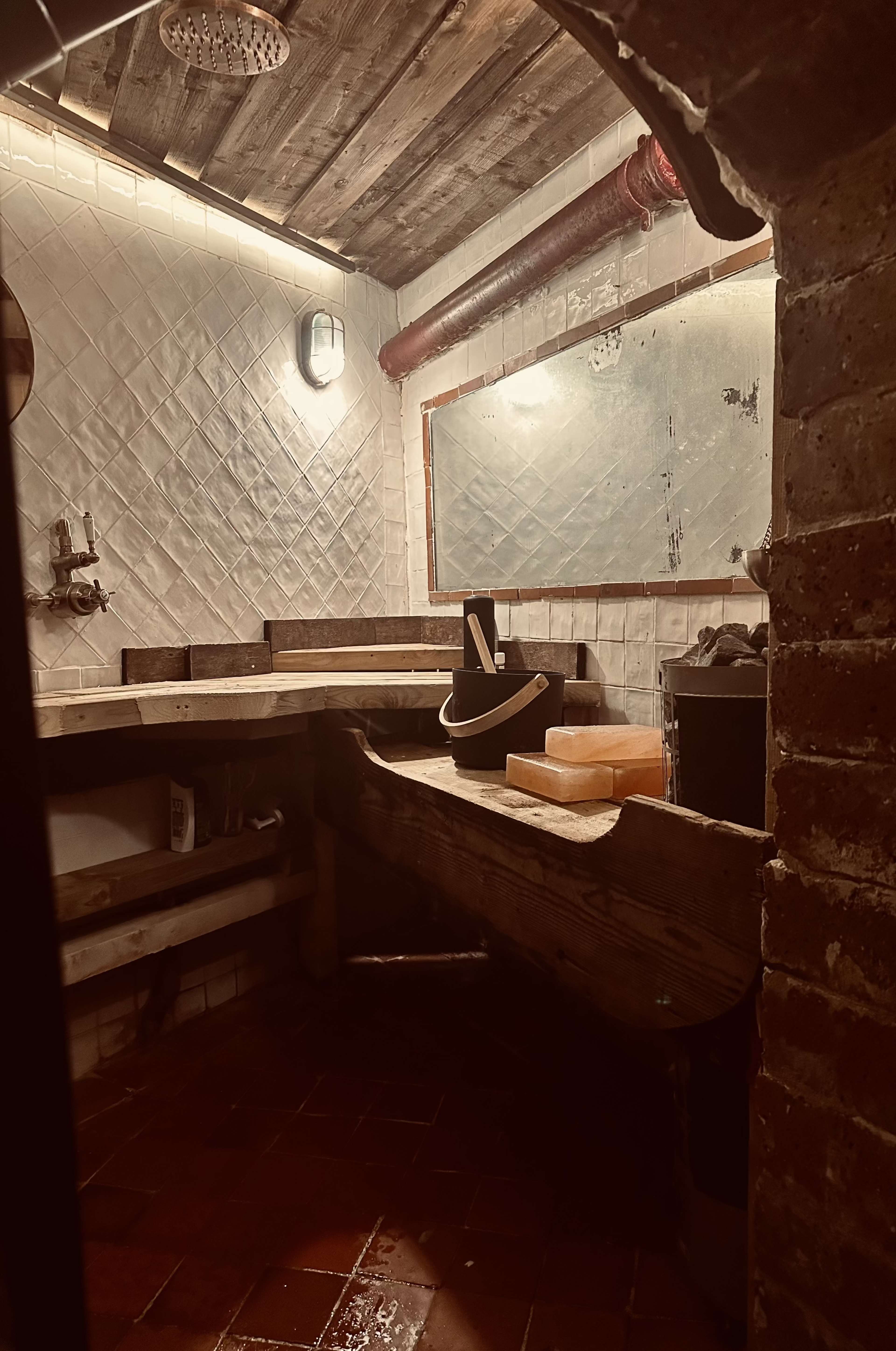A dimly lit room with wooden benches, a rustic brick wall, and a foggy mirror above a sink.