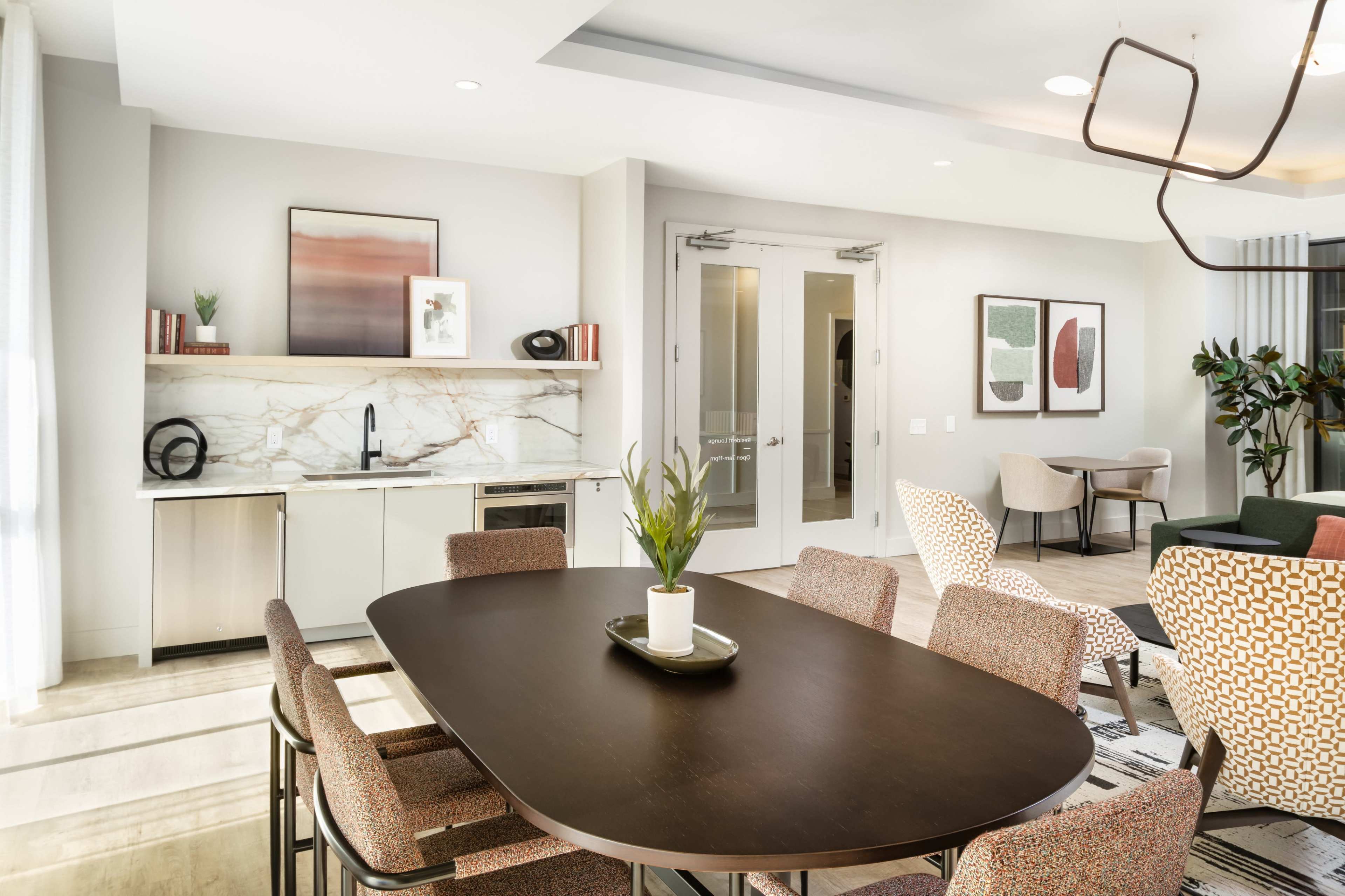 The image shows a modern dining area featuring a large table surrounded by chairs, with a kitchenette and sliding glass doors in the background.