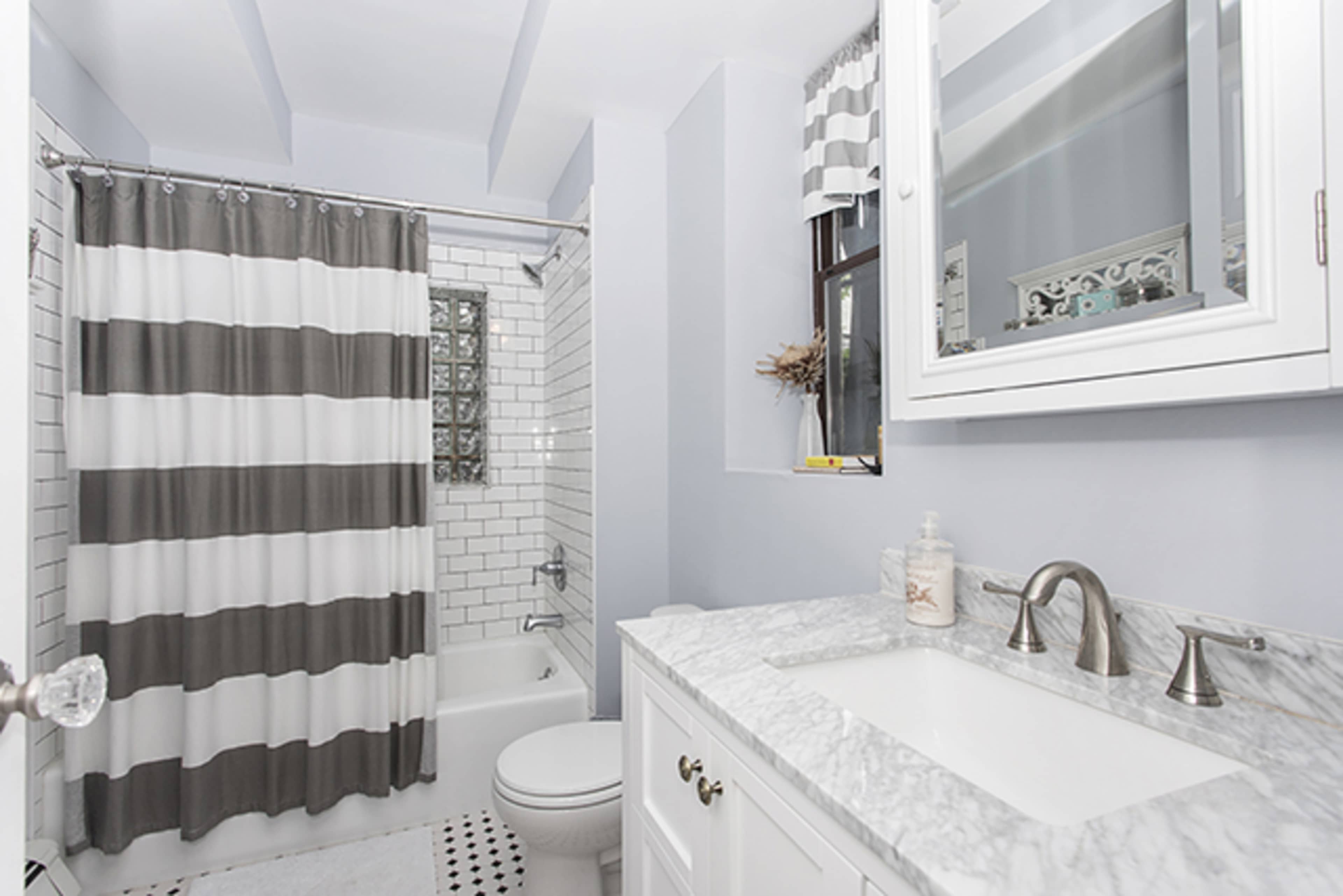 The image shows a modern bathroom featuring a white and gray striped shower curtain, a marble countertop with a sink, a toilet, and a glass-block window.
