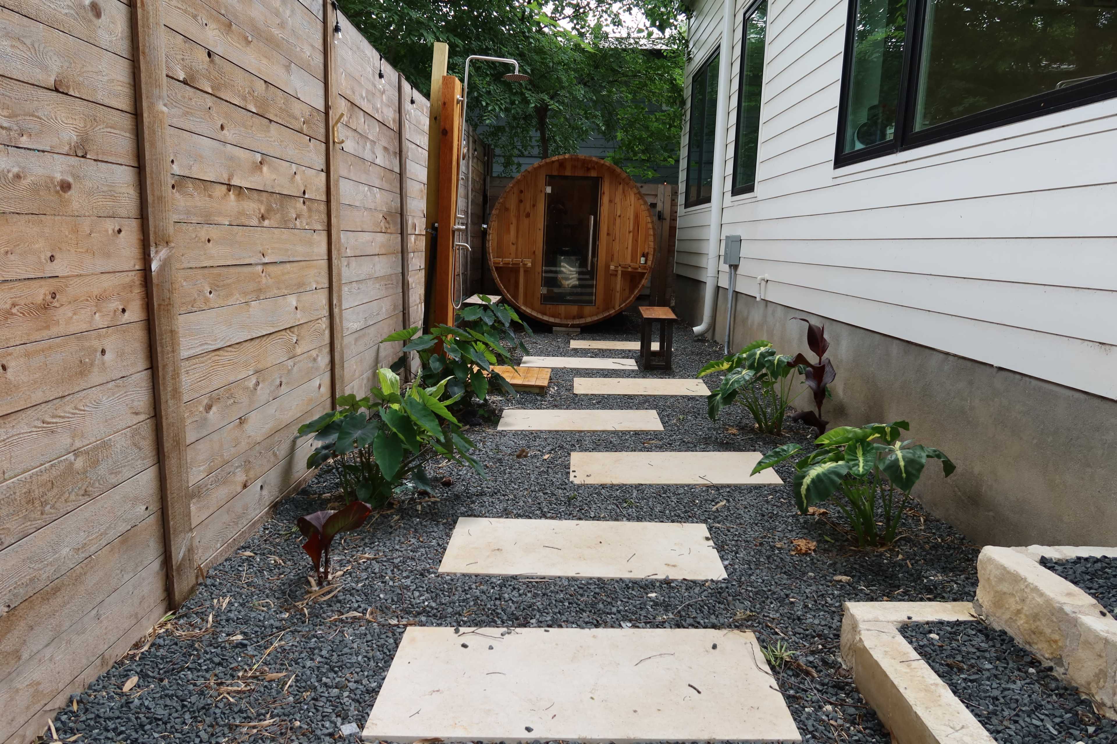 A narrow pathway lined with stone slabs and dark gravel leads to a wooden sauna, flanked by plants and wooden fencing.