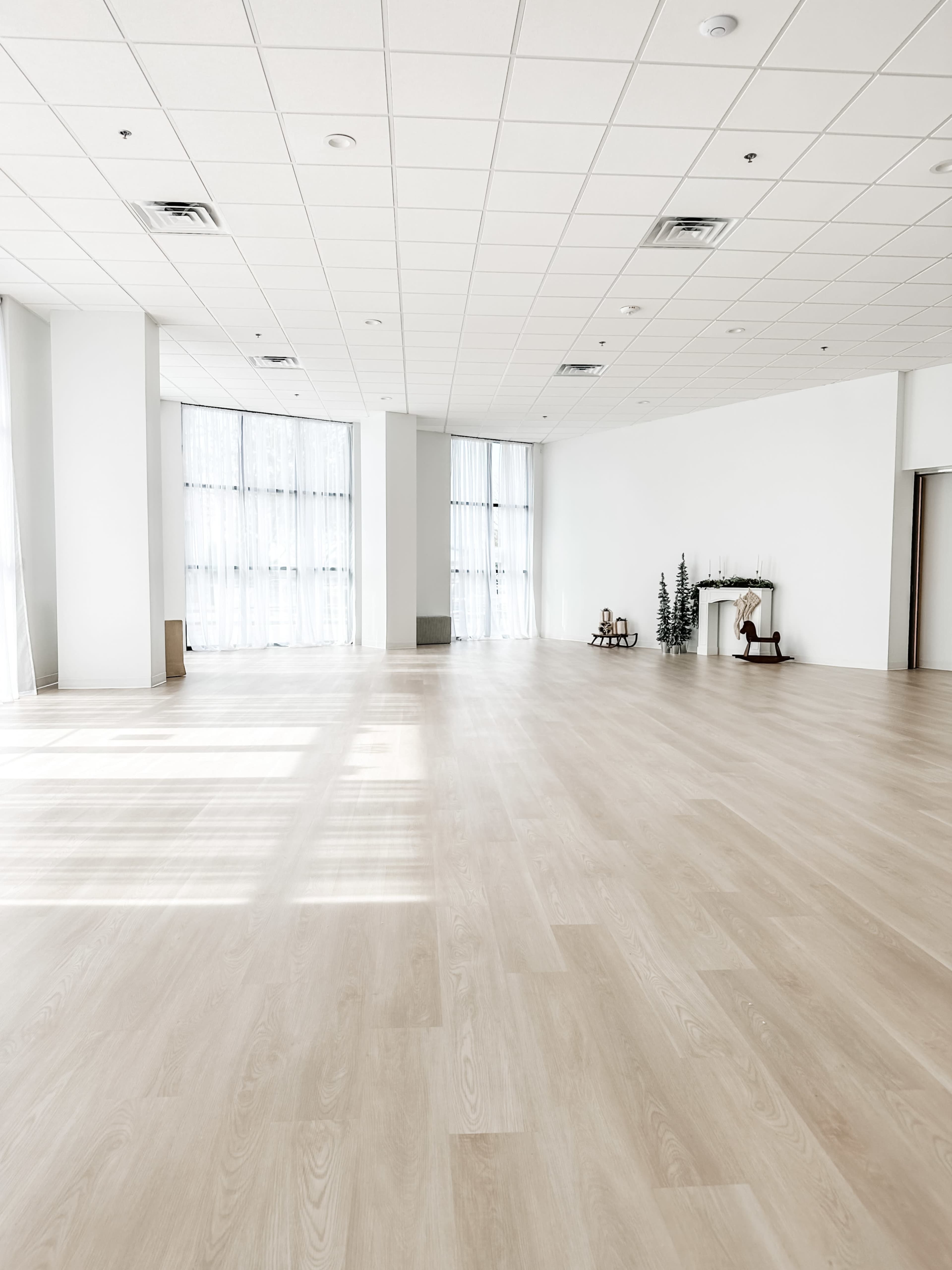 A spacious, empty room with light-colored wooden flooring and large windows allowing natural light to enter.