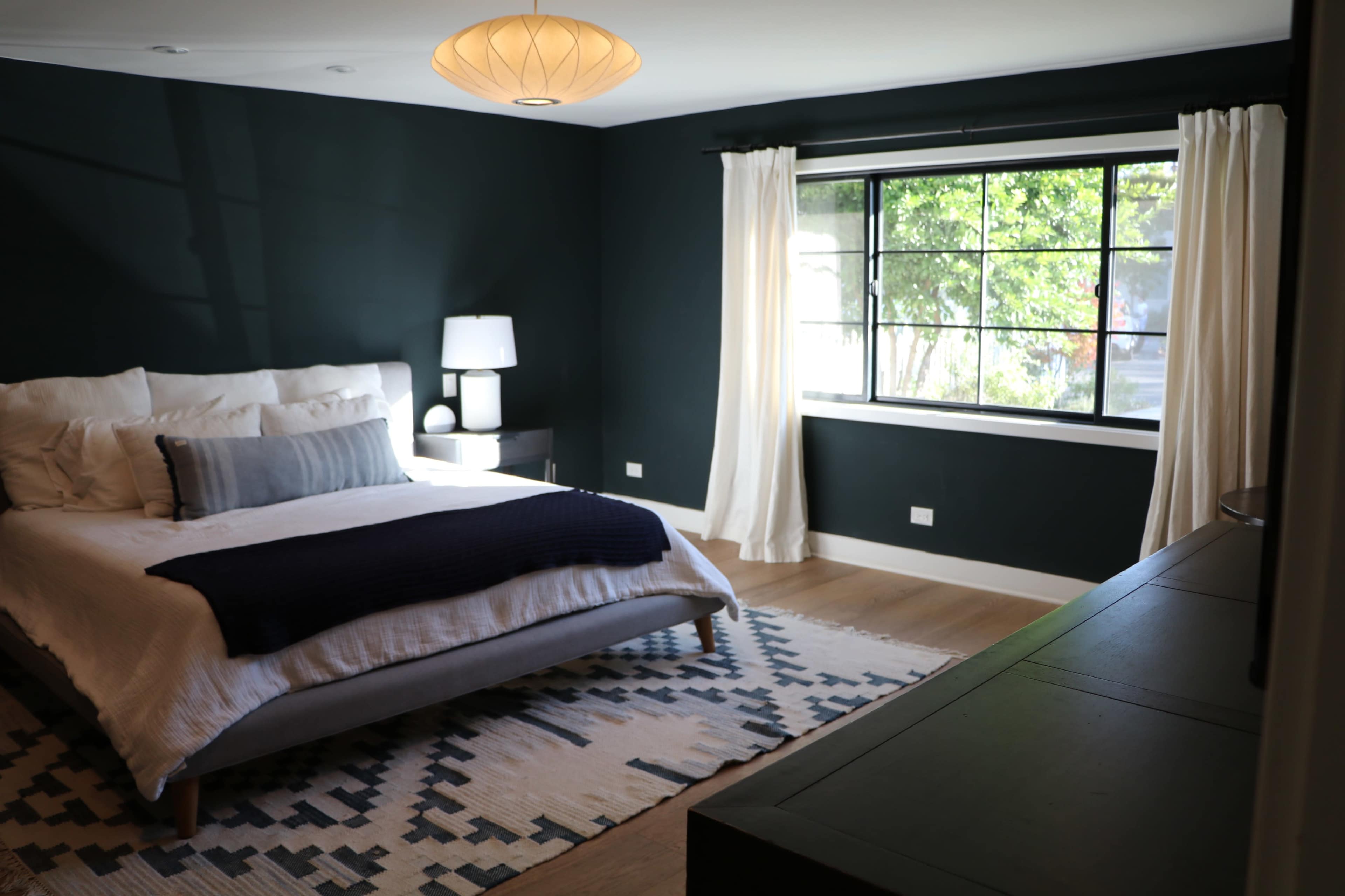 A bedroom features a bed with white linens, a dark blue throw, and a large window with sheer curtains, allowing natural light to illuminate the space.
