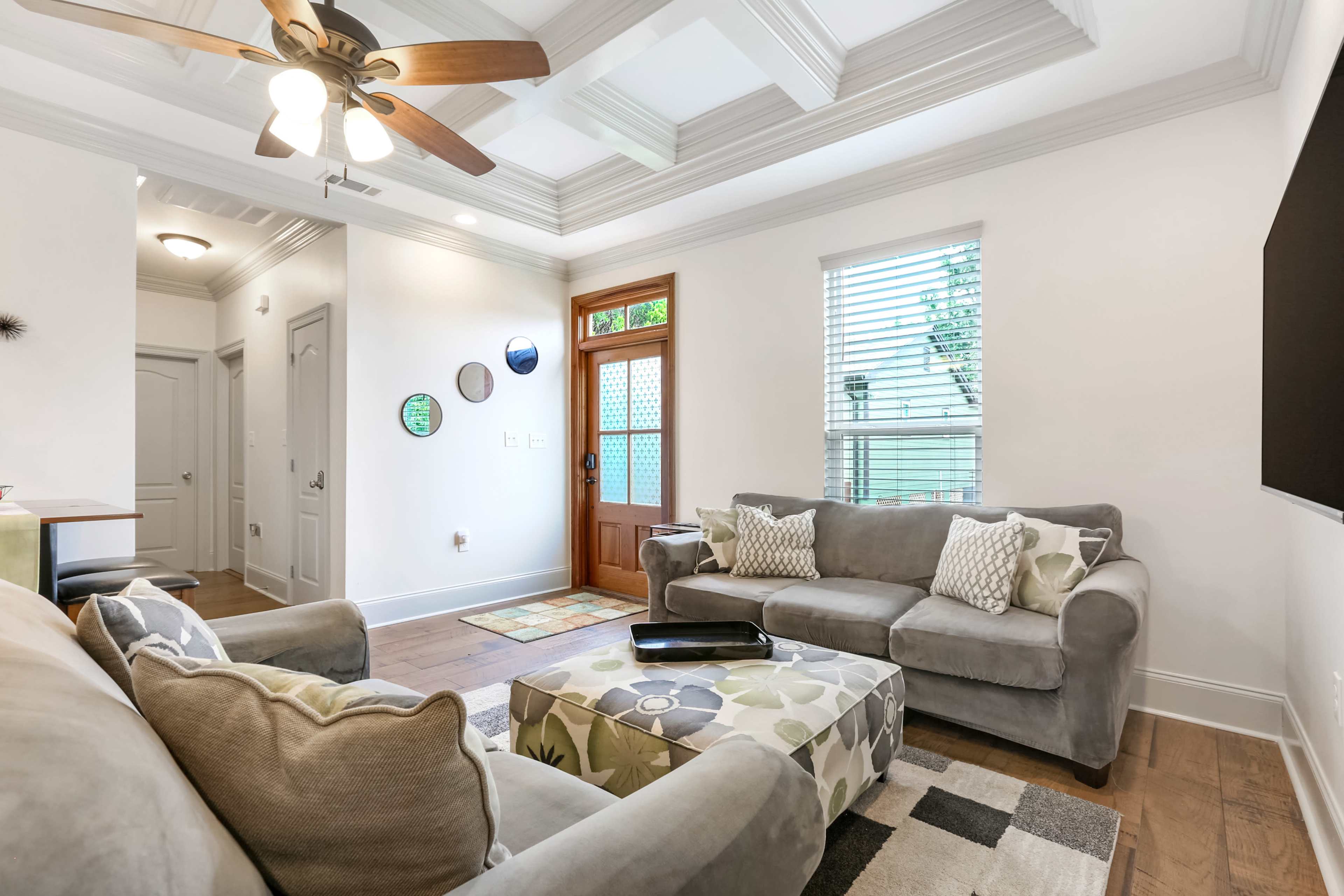 The image shows a bright living room with a ceiling fan, gray sofas, a decorative ottoman, and a doorway leading outside.
