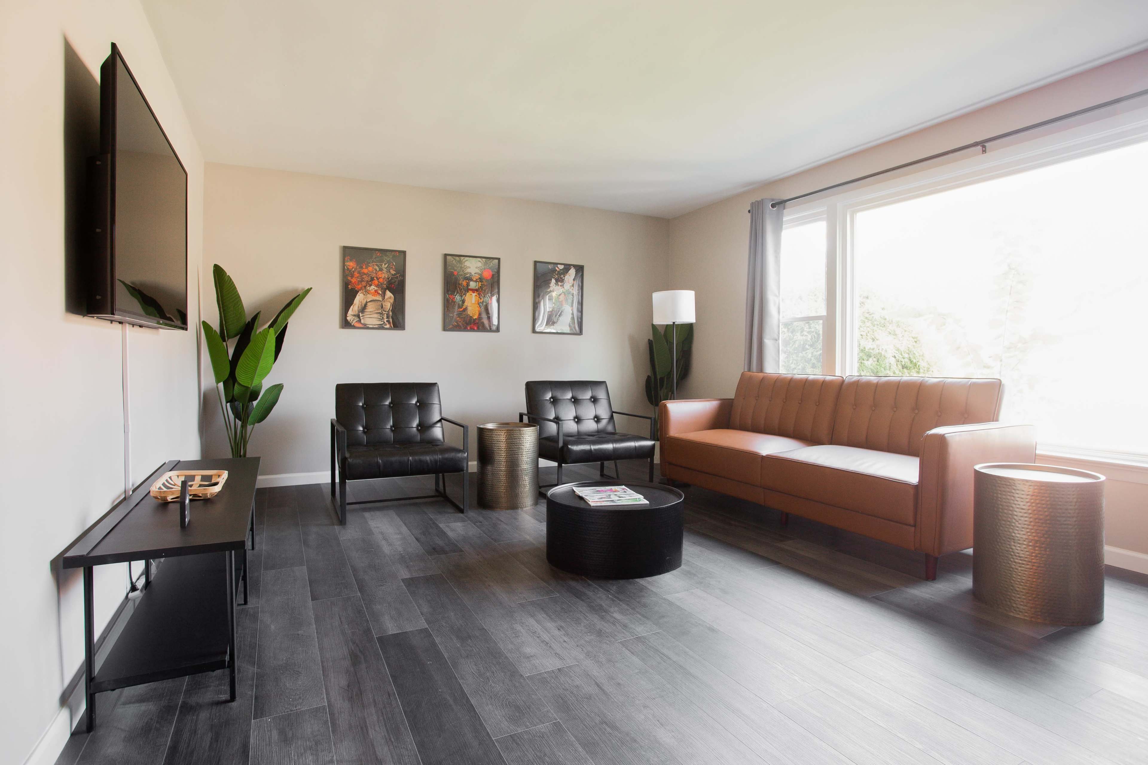 A modern living room with a brown leather sofa, two black chairs, a circular coffee table, and framed artwork on the walls, illuminated by natural light from a large window.