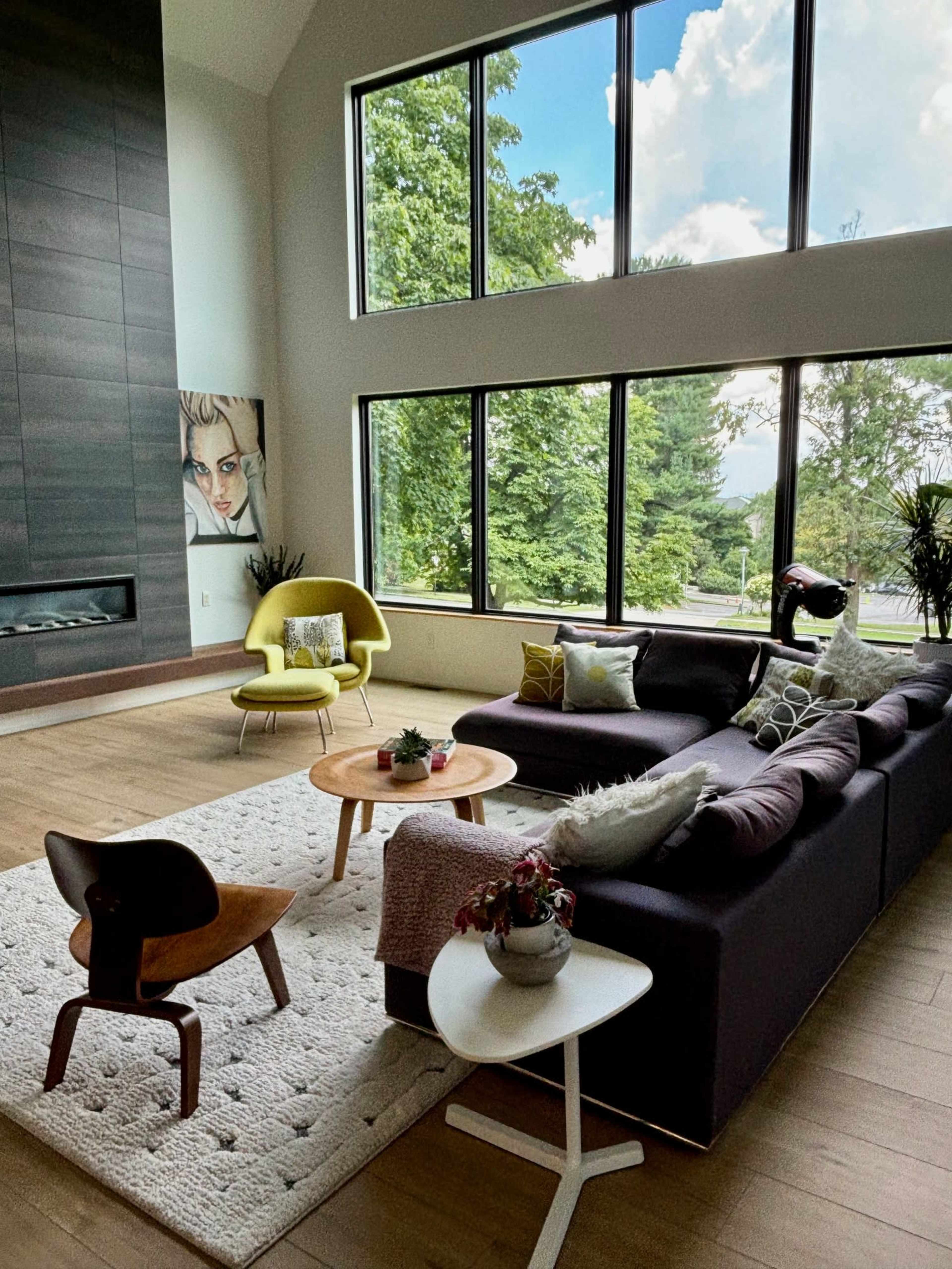A modern living room with large windows overlooking greenery, a sectional sofa, a round coffee table, and a mix of decorative plants and art.