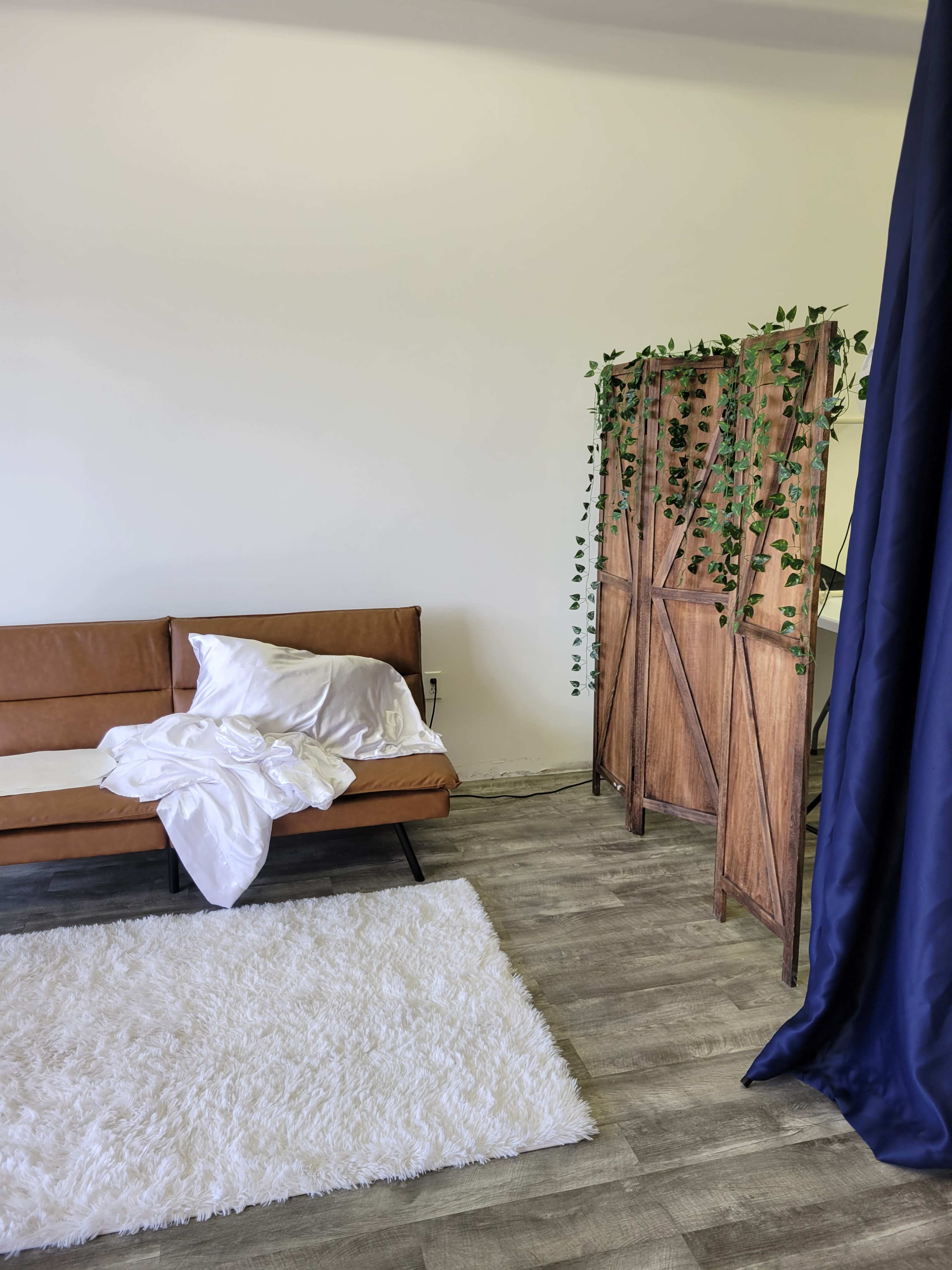 The scene features a brown sofa with rumpled white bedding and a wooden screen adorned with greenery, set against a plain wall and a plush white rug.