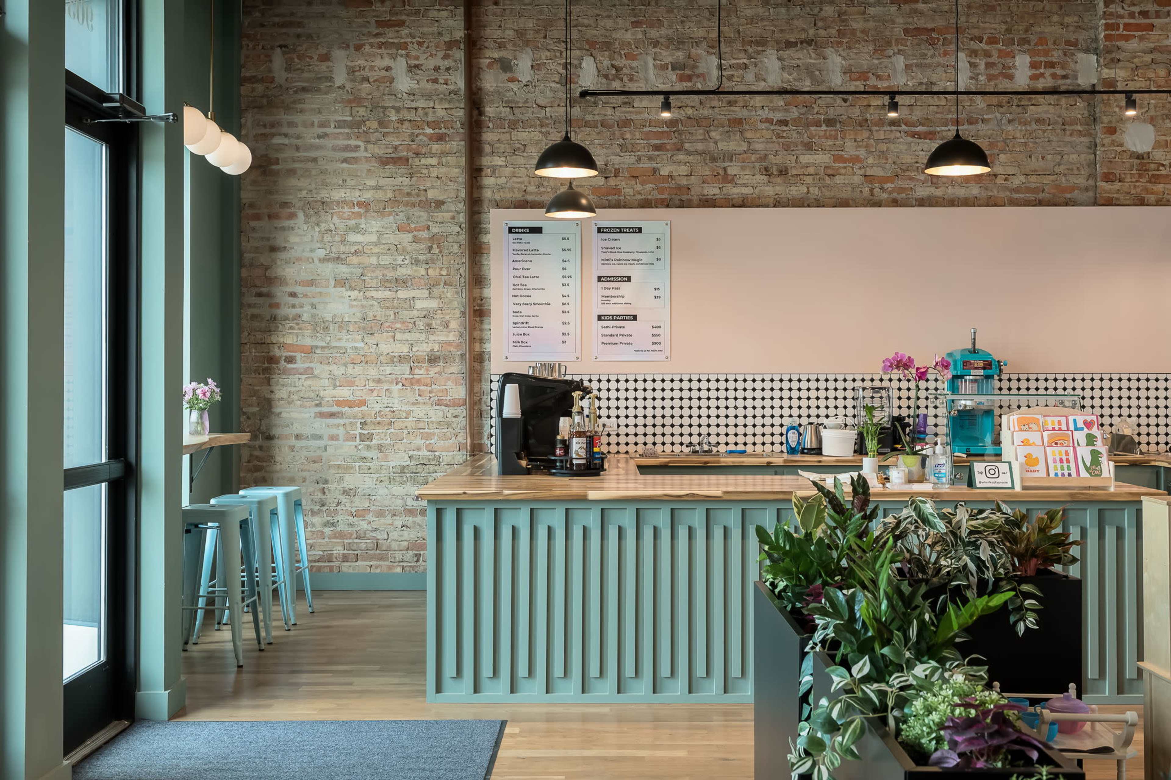 The image shows a modern café interior featuring a wooden counter with a menu board, bar stools, and potted plants against a brick wall.