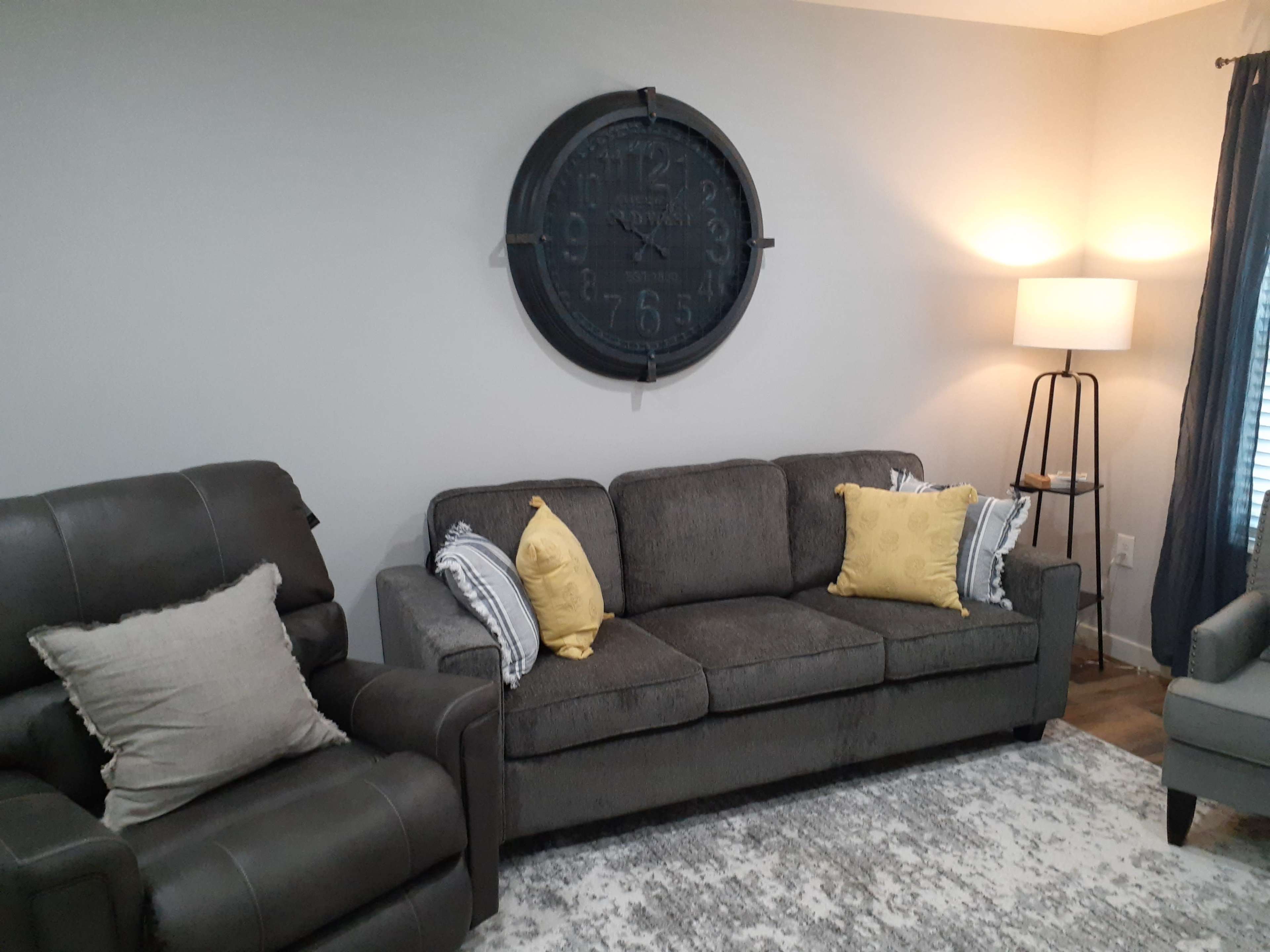 A living room with a gray couch and armchair, a round wall clock, a standing lamp, and yellow decorative pillows on the couch.