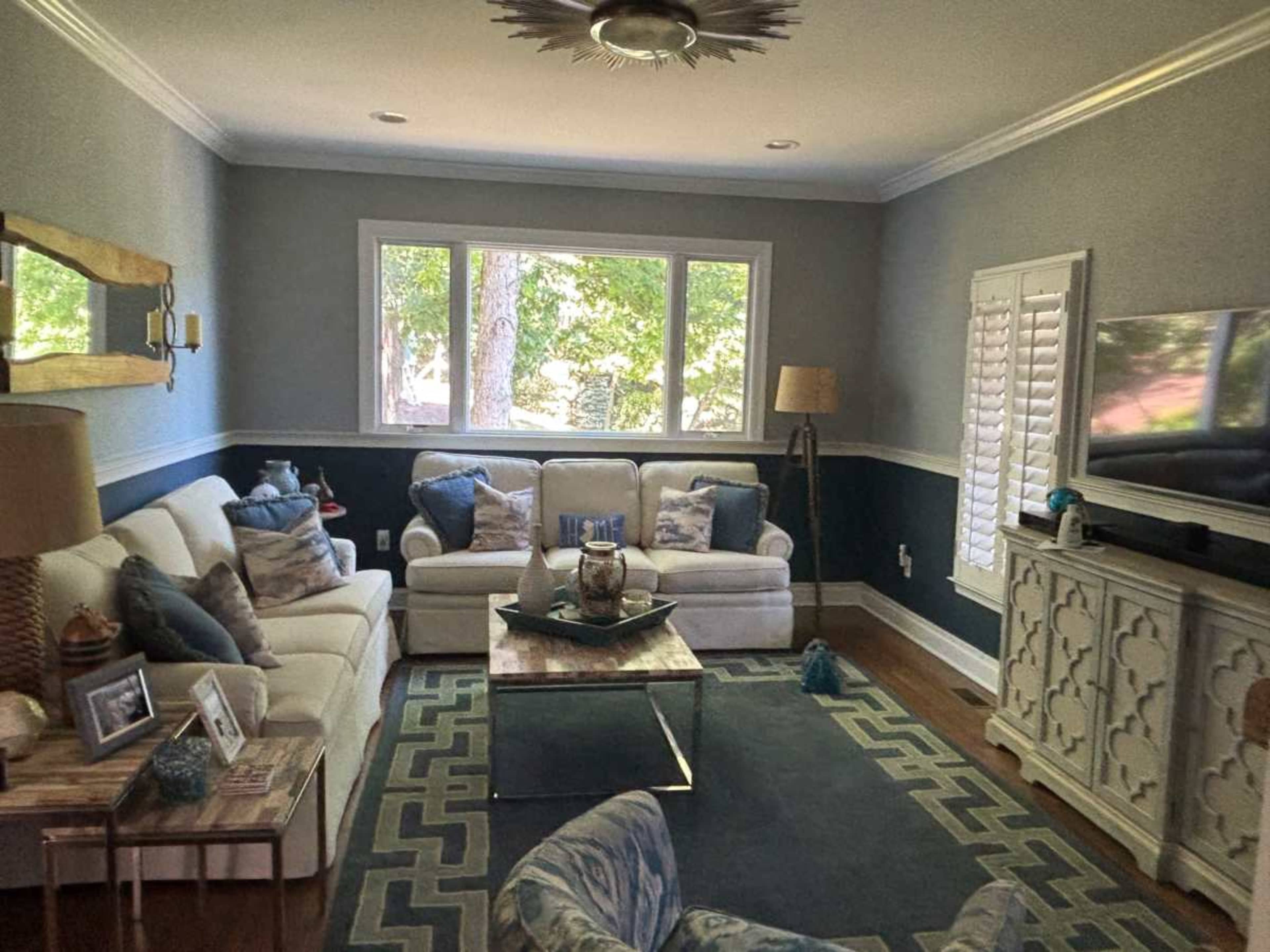 The image shows a living room with two white sofas, a coffee table, and a television, all arranged around a patterned area rug under natural light from large windows.