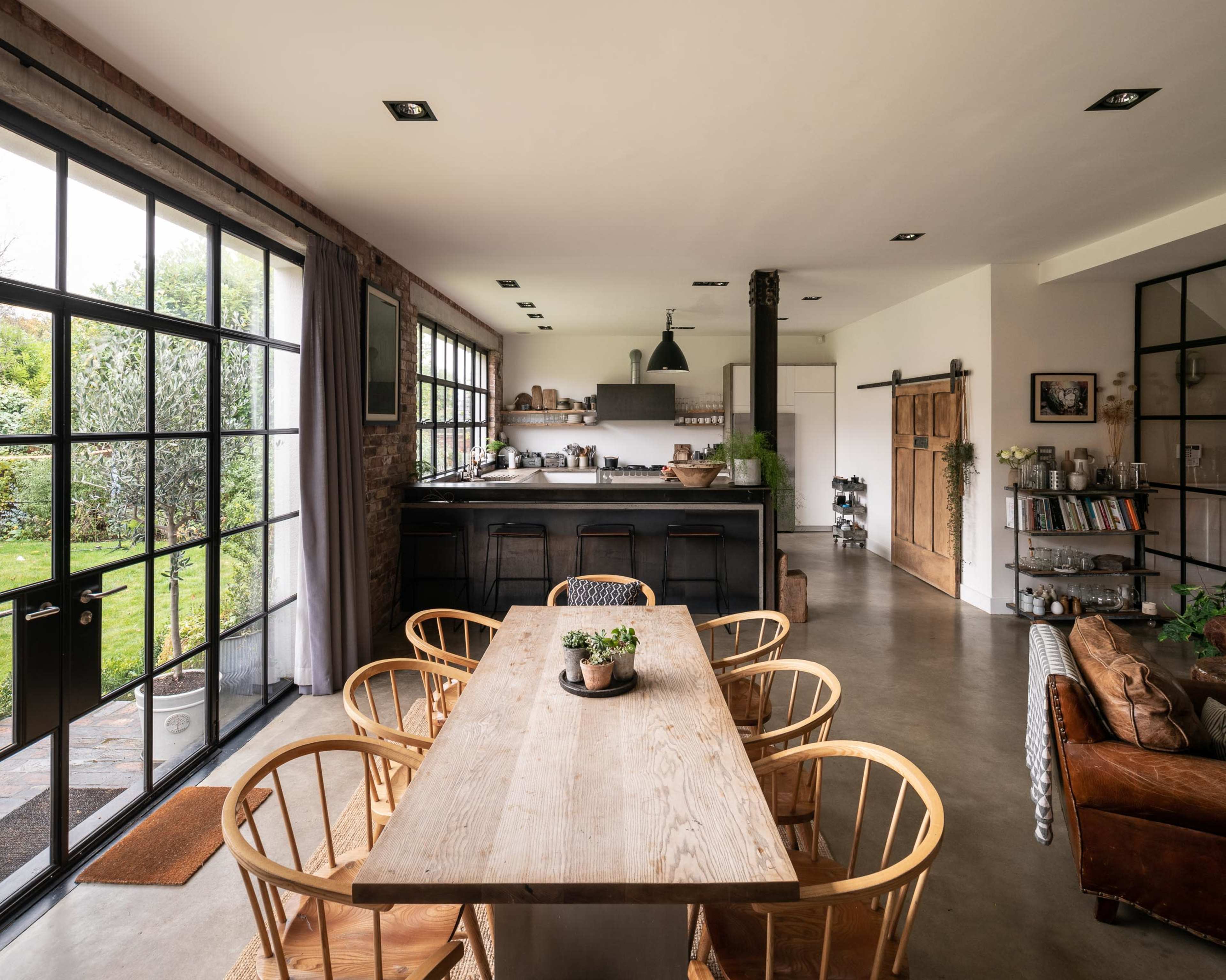 A modern kitchen and dining area with a large wooden table, surrounding chairs, and large windows that open to a garden.