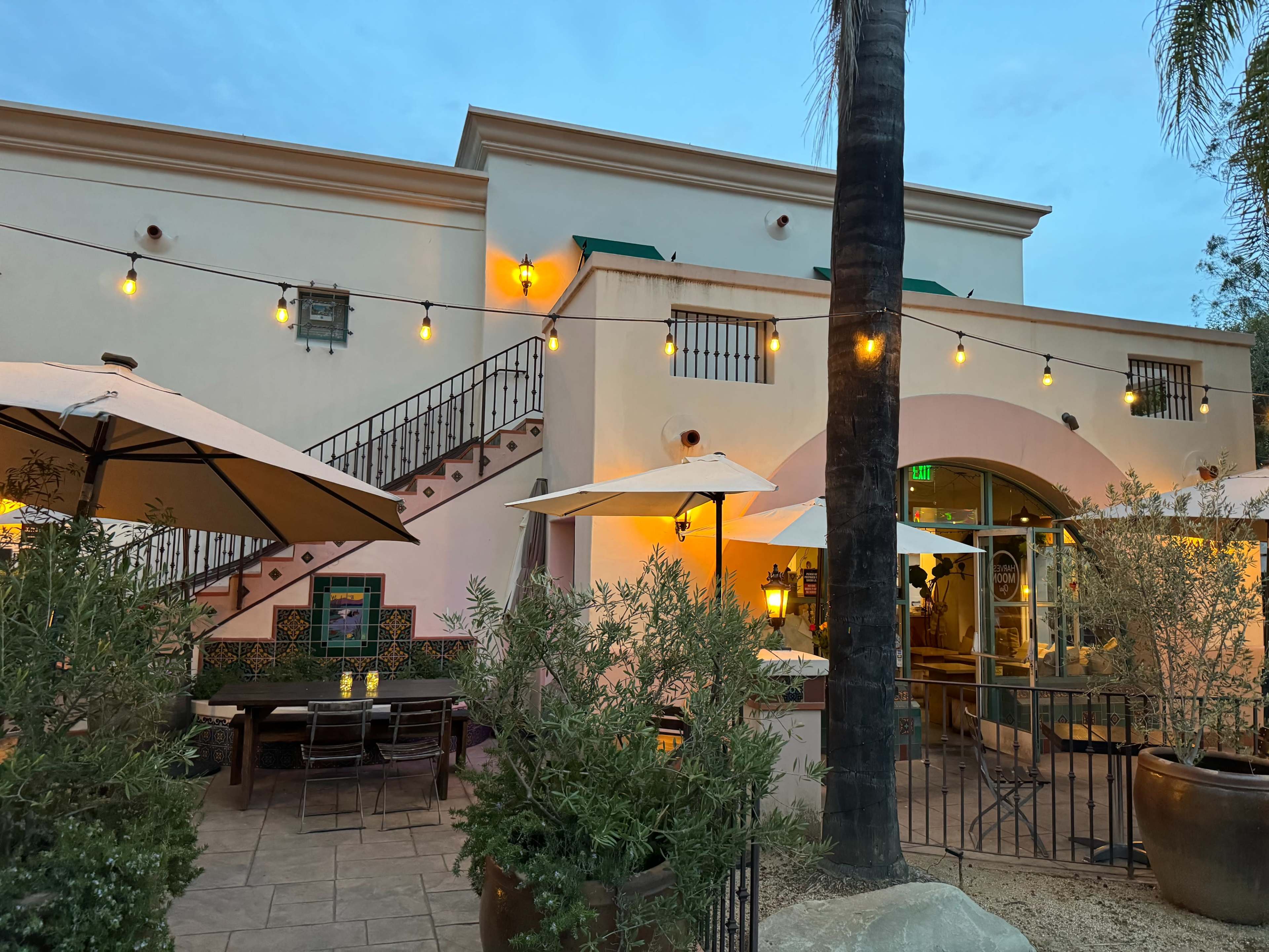 A restaurant patio with string lights, umbrellas, and an outdoor dining area adjacent to a building with stairs and a large archway.