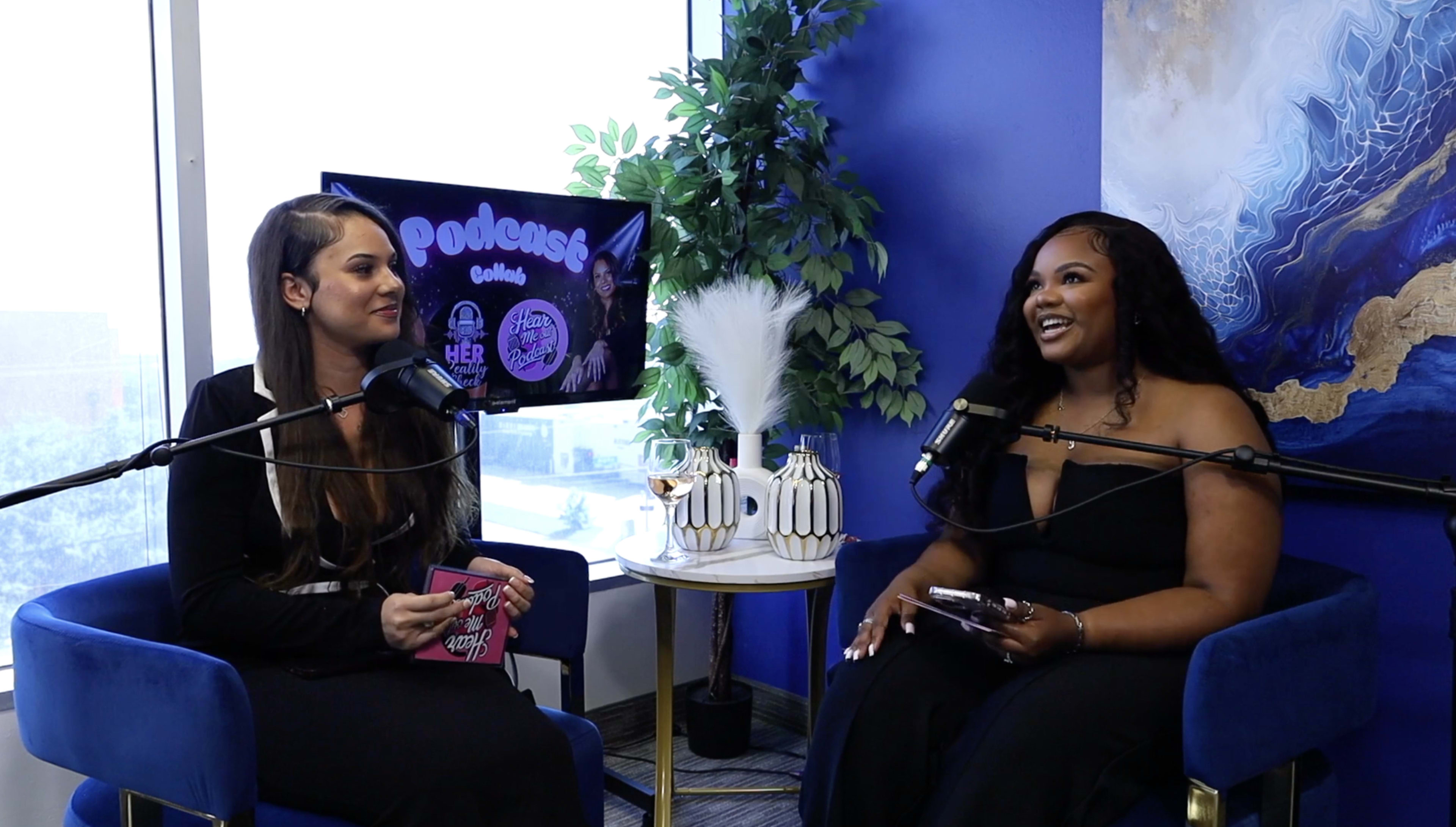 Two women are sitting in chairs with microphones, engaging in a conversation in a well-lit room adorned with plants and decorative elements, while a television displays podcast branding in the background.