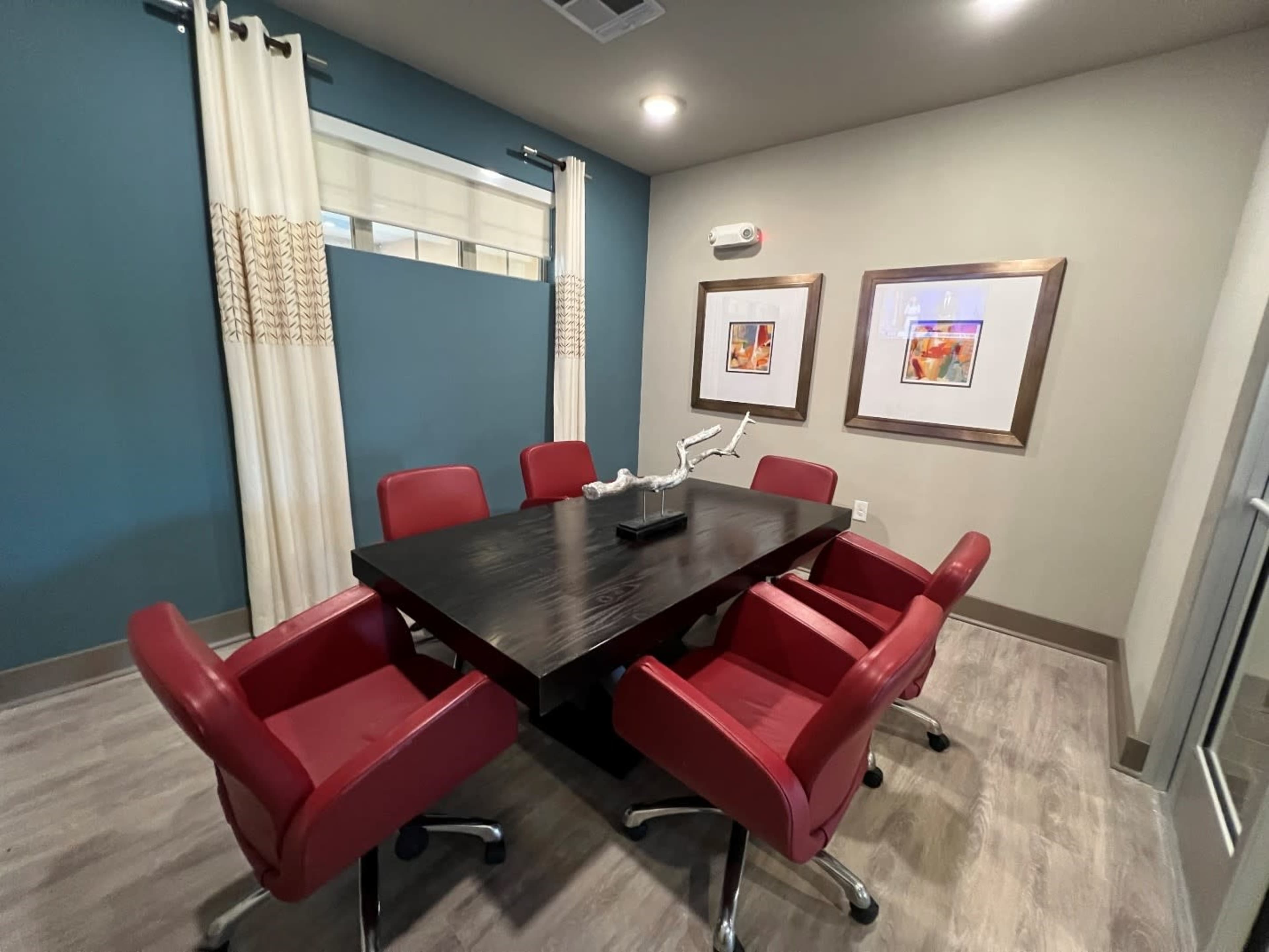 A conference room features a large black table surrounded by red chairs, with artwork hanging on the wall and curtains framing a window.