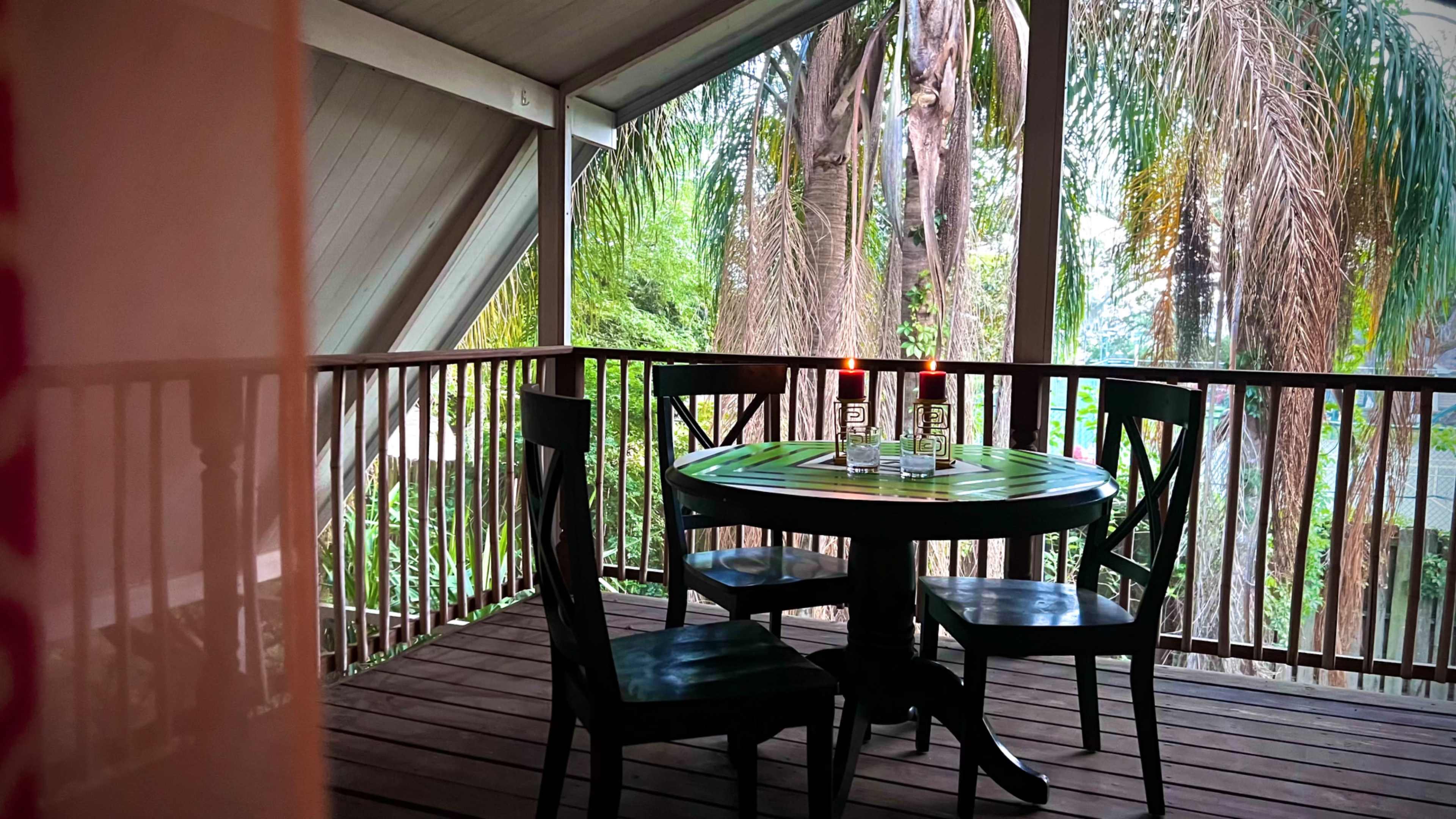 A small wooden dining table with four chairs is set on a porch surrounded by tropical greenery.