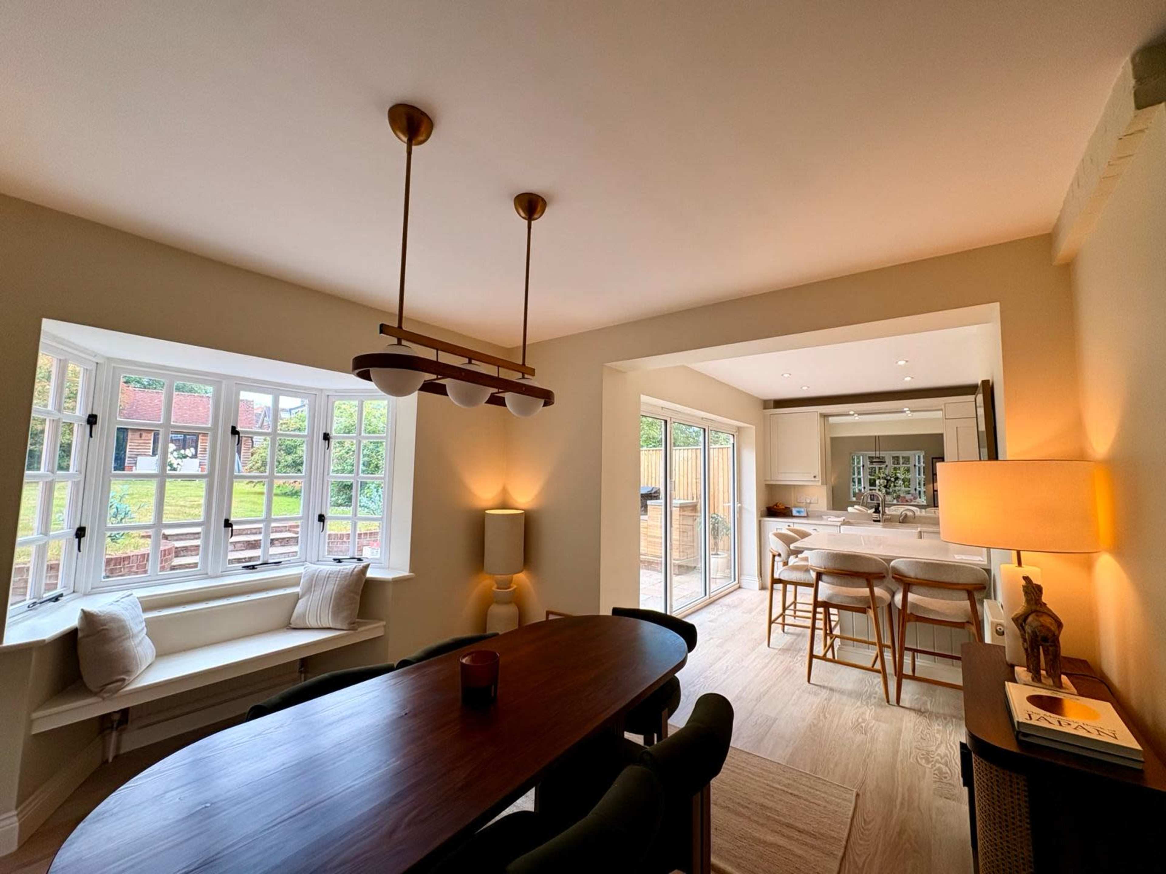 A dining area features a long table with a chandelier overhead, and sunlight streams in through large windows, leading into a modern kitchen visible in the background.