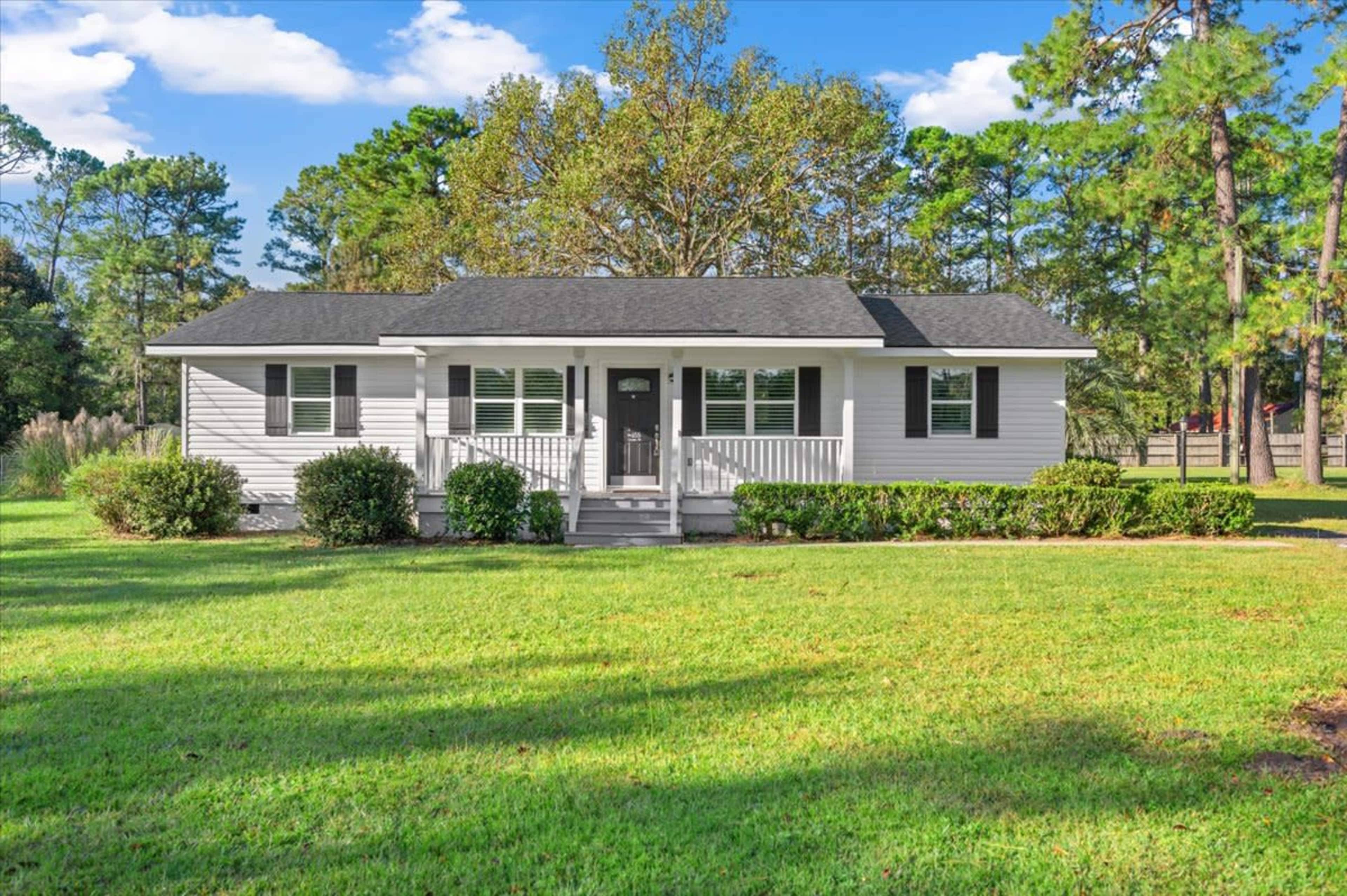 A single-story house with a black roof and white siding sits in a grassy yard surrounded by trees.